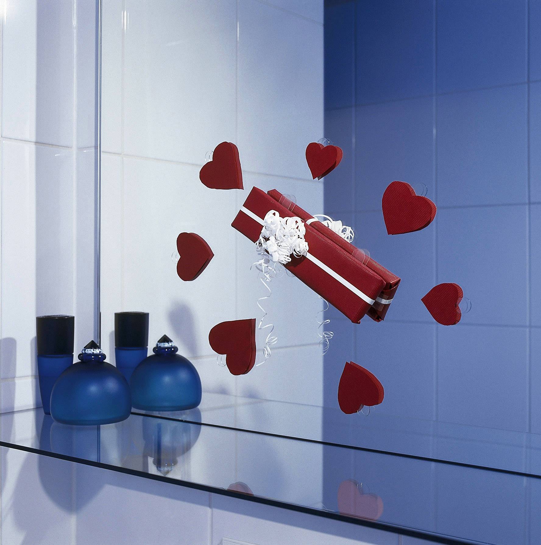 Cadeau rouge avec des rubans blancs flottant, entouré de cœurs rouges dans une pièce aux carreaux bleus, à côté de bouteilles bleues.