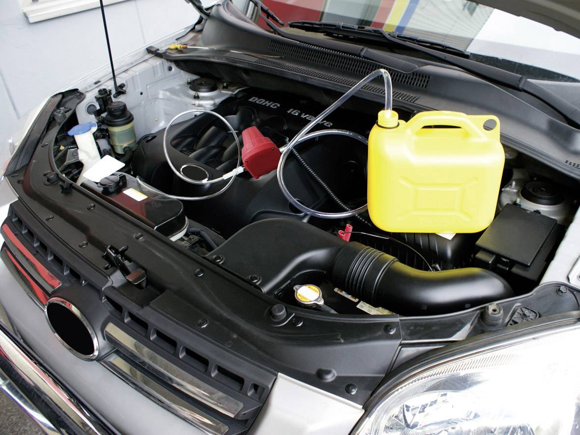 Un compartiment moteur ouvert avec un bidon jaune posé sur le bloc moteur et des tuyaux connectés au bidon, illustrant un système de remplissage de liquide de refroidissement.