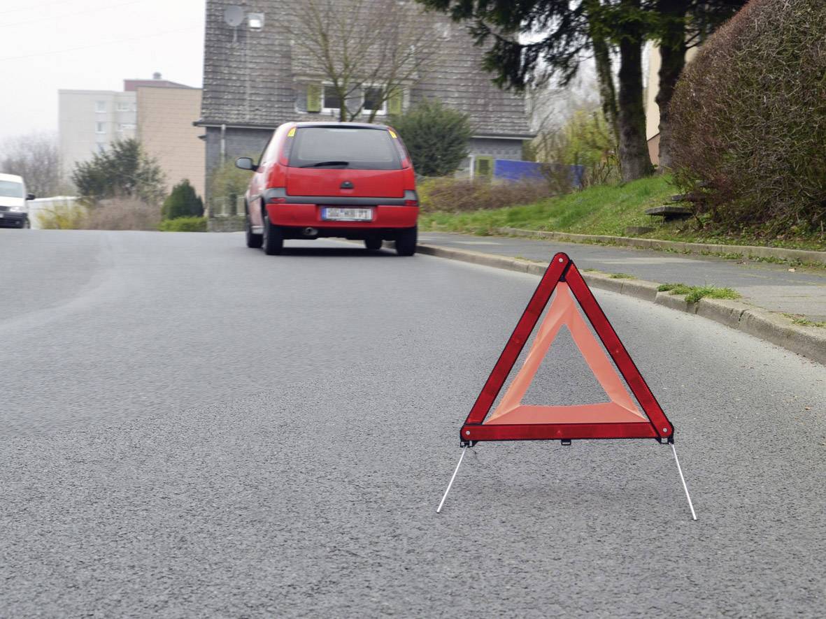Un triangle de signalisation est placé sur une route, tandis qu'une voiture rouge est garée en arrière-plan. La scène suggère une panne potentielle.