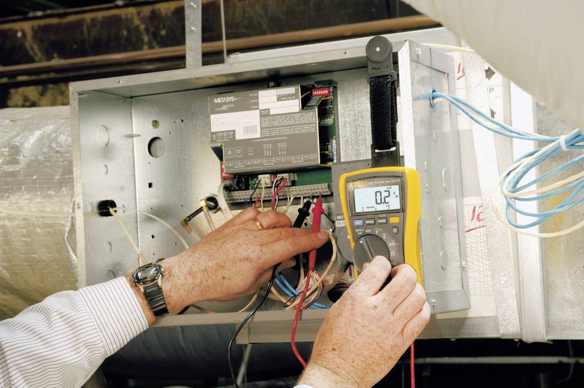 Une personne mesure l'électricité d'un système de ventilation à l'aide d'un multimètre jaune affichant '0.2' et vérifie les connexions de câbles.