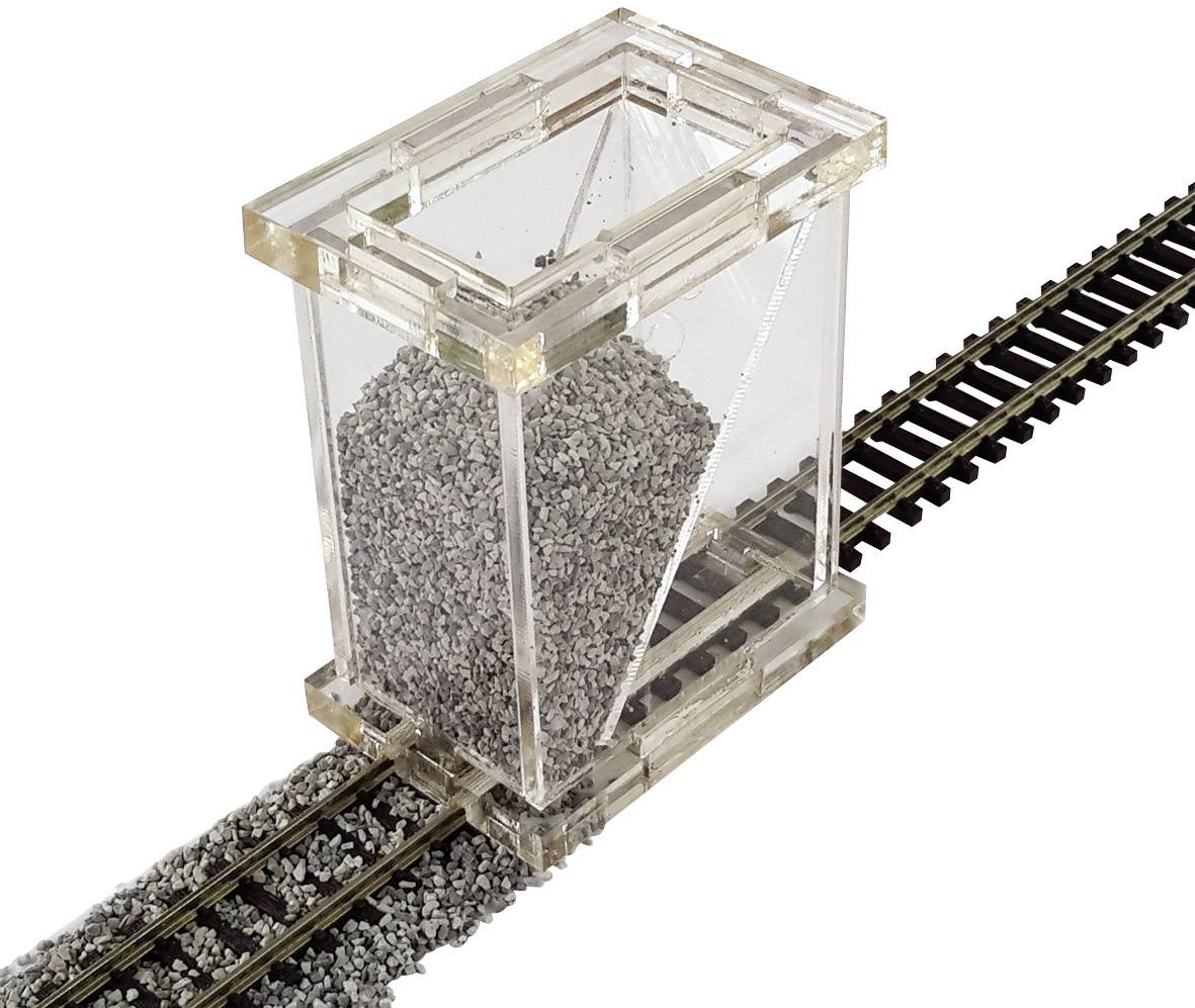 Un récipient transparent rempli de galets gris est posé sur un rail de train miniature, avec quelques pierres dispersées sur le rail.