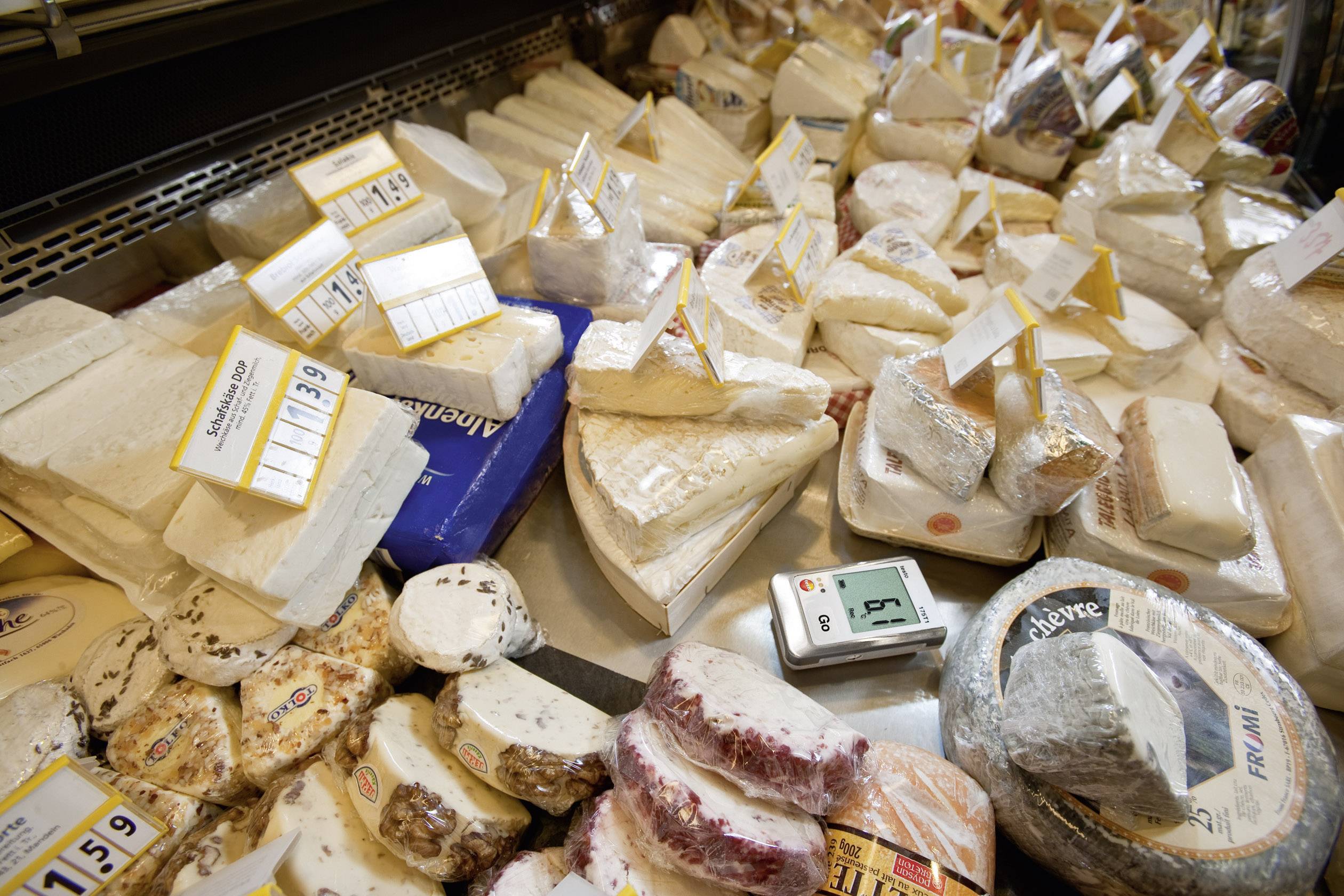 Différentes sortes de fromages sont exposées sur un étal dans un supermarché, avec des étiquettes indiquant le prix et le poids de chaque pièce.