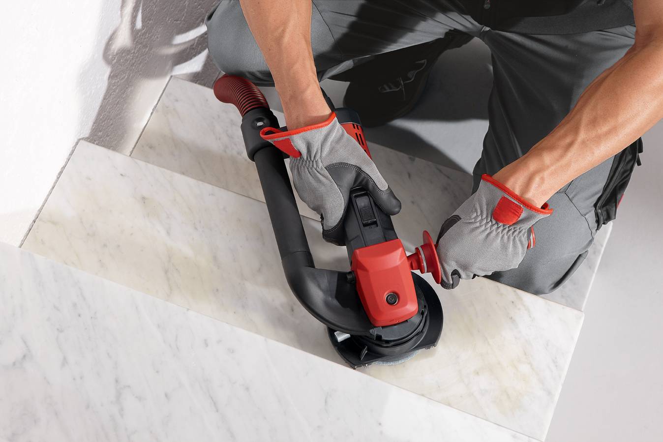 Une personne coupe quelque chose avec une meuleuse d'angle sur un escalier en marbre. Elle porte des gants gris et des vêtements de travail.
