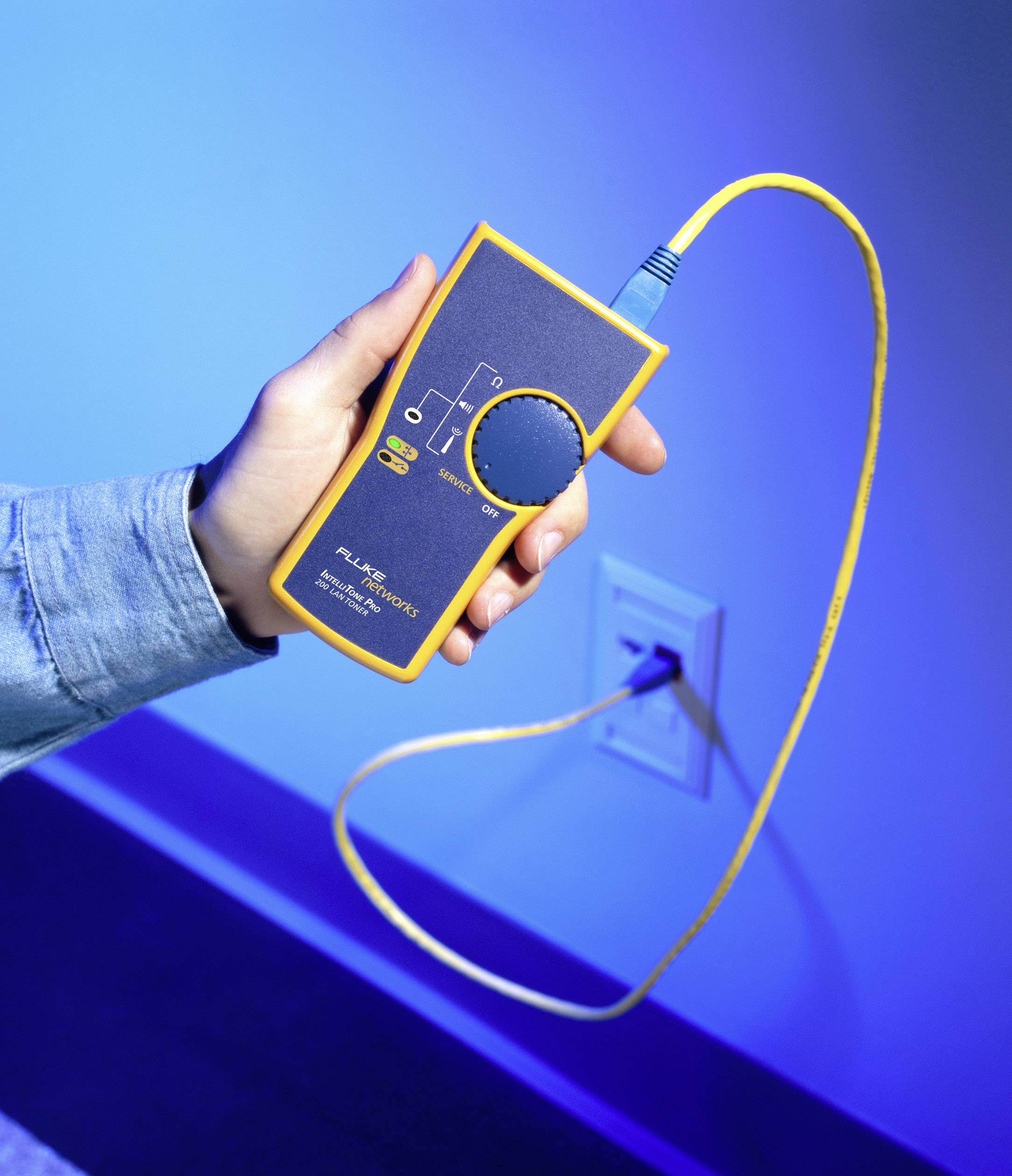Une personne tient un appareil de test de câble réseau jaune devant un mur bleu. L'appareil sert à diagnostiquer les connexions réseau.