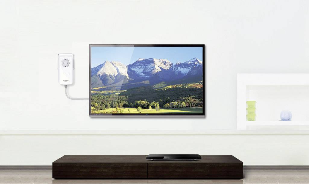 Un téléviseur à écran plat est accroché à un mur blanc, affichant un paysage de montagnes et de forêts. En dessous, un meuble TV marron est presque vide, ne portant qu'un seul appareil.