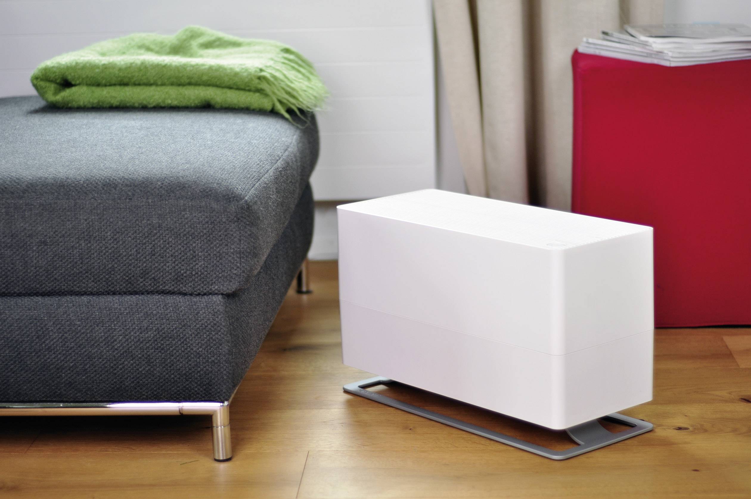 Un humidificateur blanc est posé sur un sol en bois à côté d'un canapé gris avec une couverture verte. Un tabouret rouge se trouve en arrière-plan.