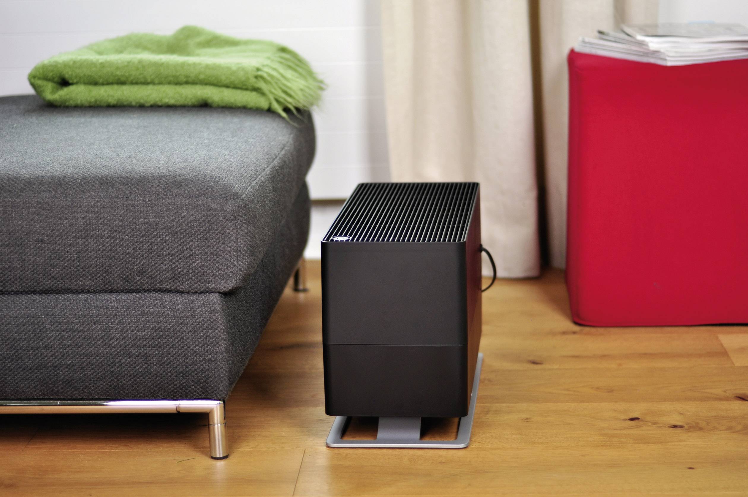 Un humidificateur moderne est posé sur un sol en bois, à côté d'un canapé gris recouvert d'un tissu vert et d'un tabouret rouge sur lequel sont posés des journaux.