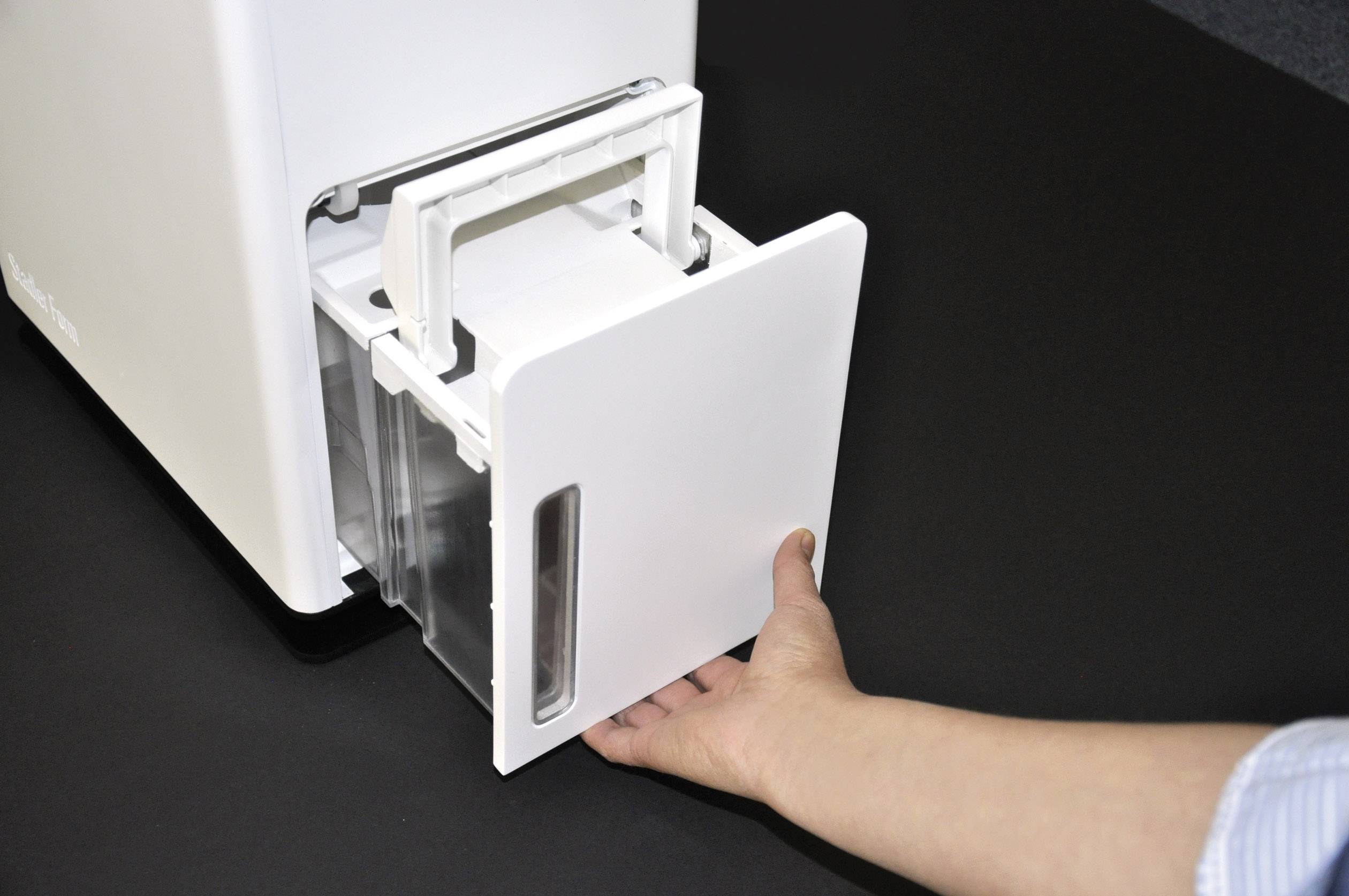 Une main sort un récipient d'un appareil blanc. Le récipient a une fenêtre transparente à l'avant et semble facile à retirer.