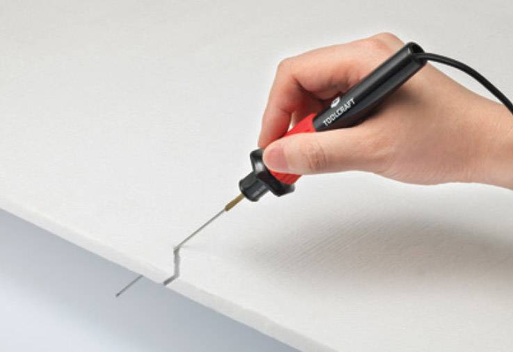 Une main tient un coupe-fil chaud, qui coupe précisément à travers un matériau blanc, probablement de la mousse ou du plastique.