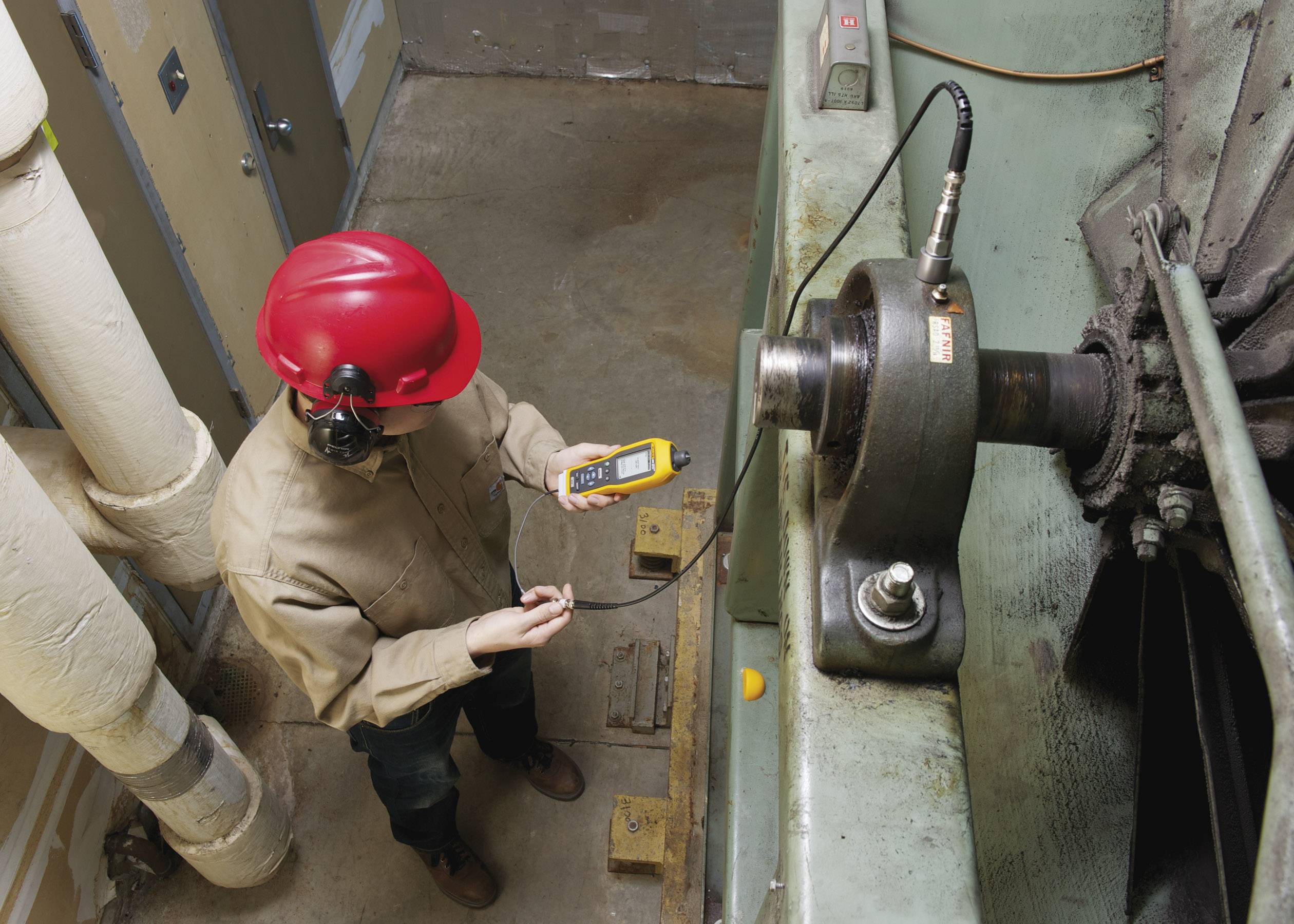 Un technicien portant un casque de protection rouge mesure les vibrations d'une machine industrielle à l'aide d'un appareil de mesure afin de vérifier son état.