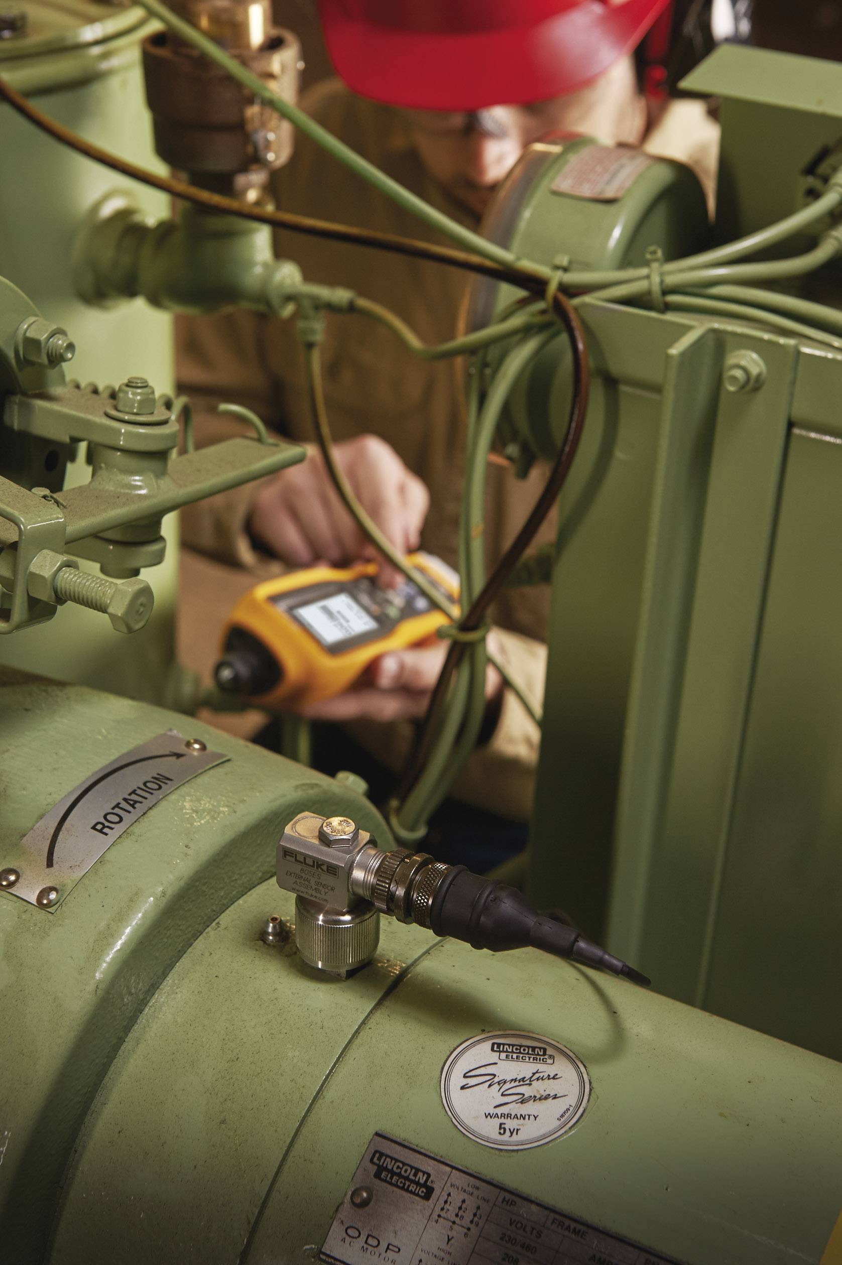 Une personne portant un casque rouge vérifie l'état d'une machine industrielle verte à l'aide d'un appareil de mesure. Des câbles sont visiblement connectés.