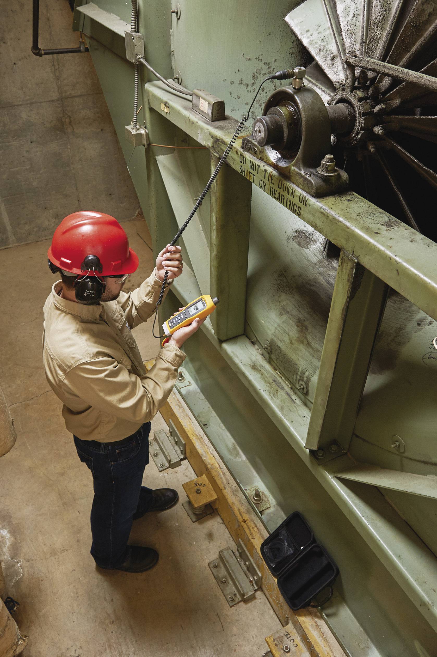 Un technicien portant un casque rouge mesure les vibrations d'une grande machine industrielle à l'aide d'un appareil.