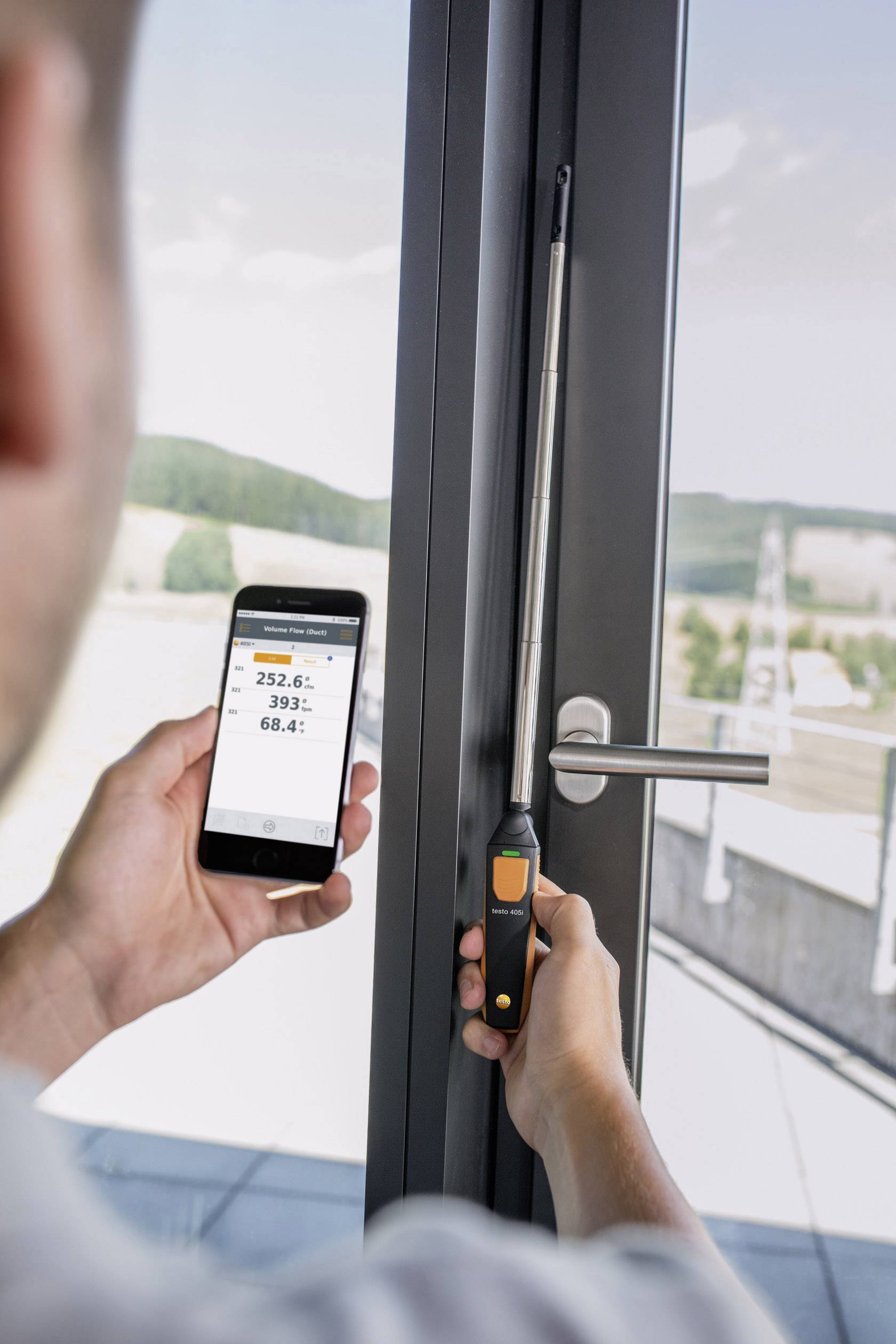 Une personne mesure l'humidité d'une fenêtre à l'aide d'un appareil qui transmet les valeurs à un smartphone. Arrière-plan : paysage.