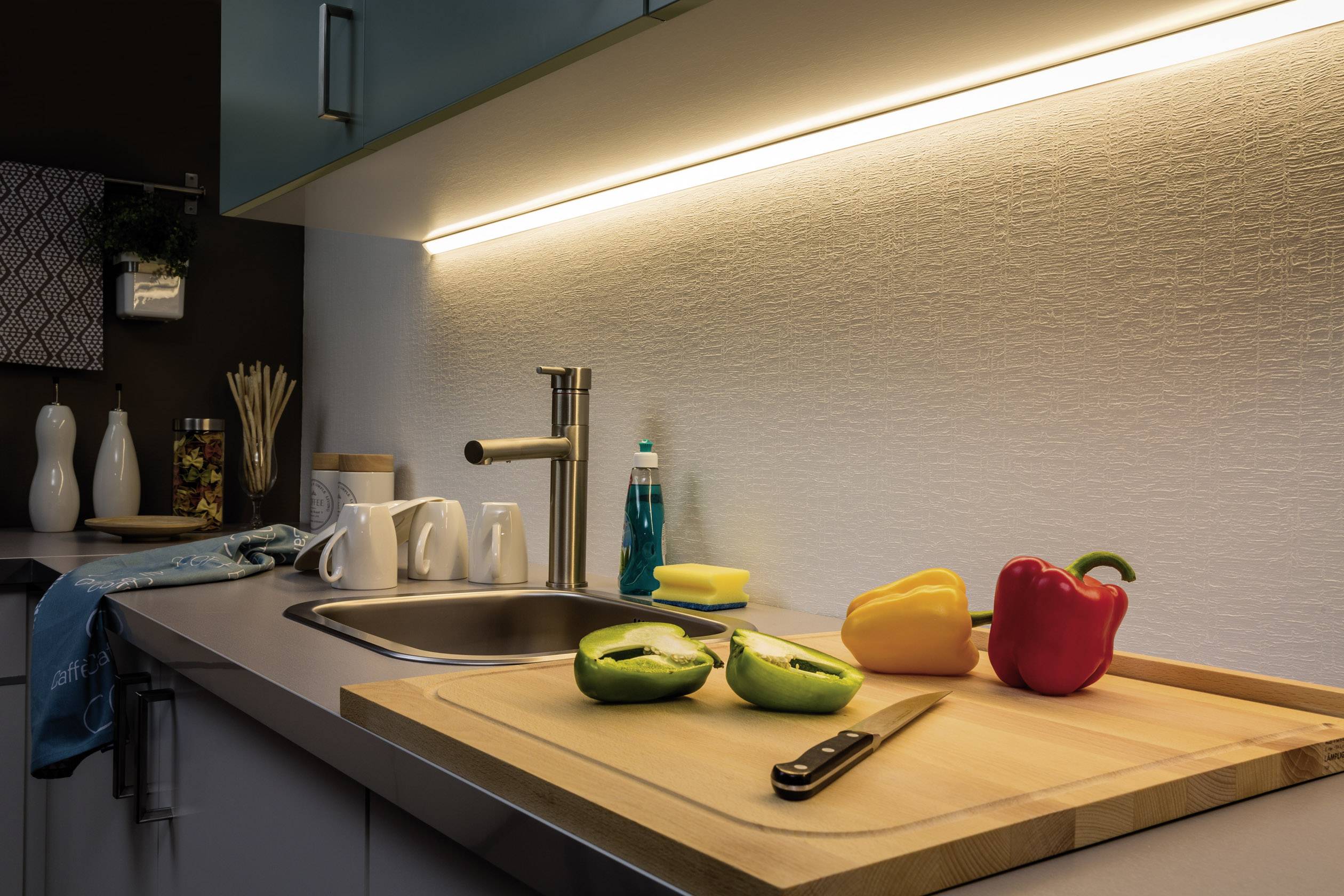 Îlot de cuisine avec espace de travail éclairé, sur lequel se trouve une planche à découper avec un couteau, un poivron vert, un poivron jaune et un poivron rouge.