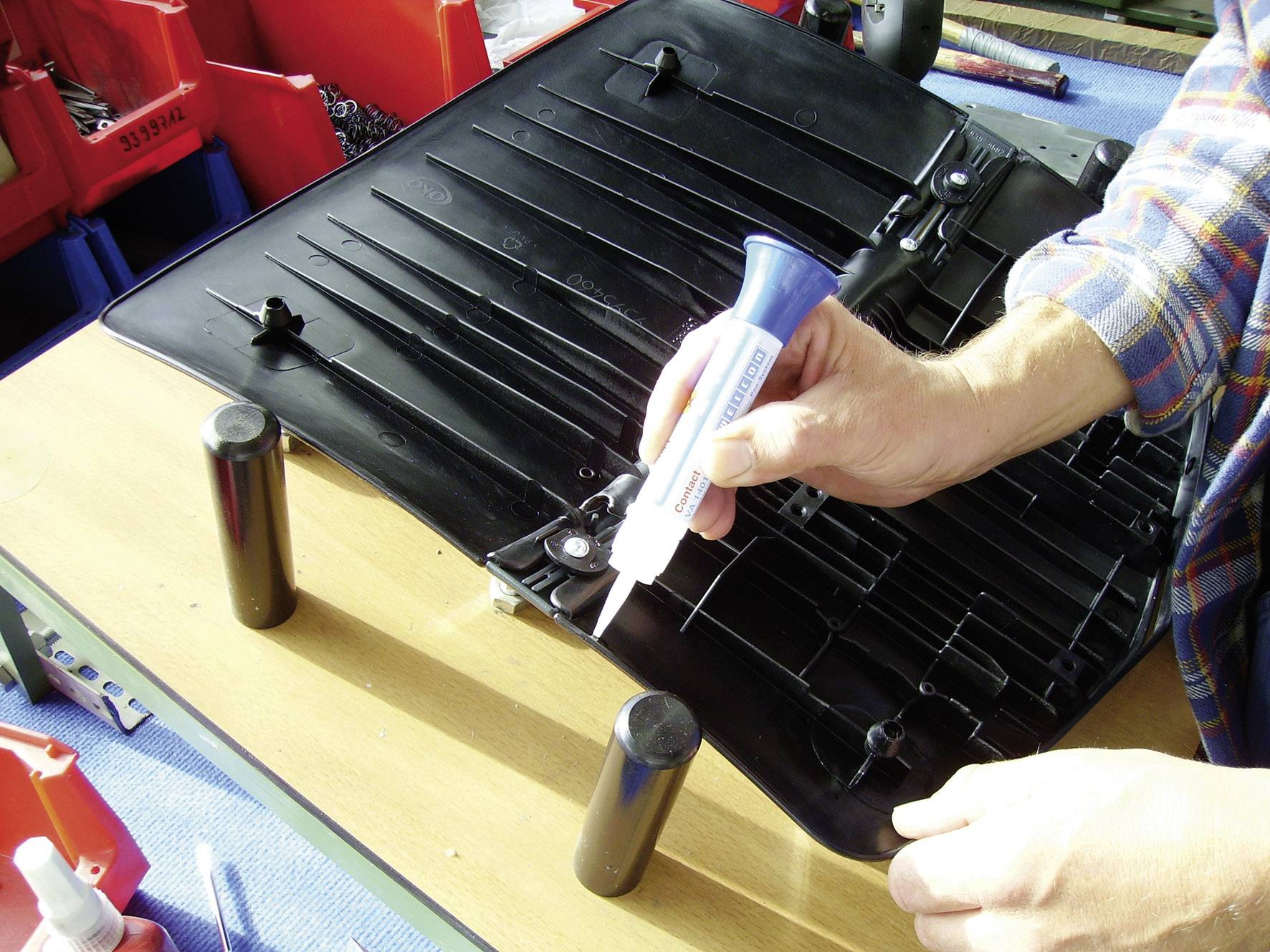 Une personne applique de la colle sur une protection en plastique noir. À l'arrière-plan, on peut voir des boîtes rouges contenant des outils.