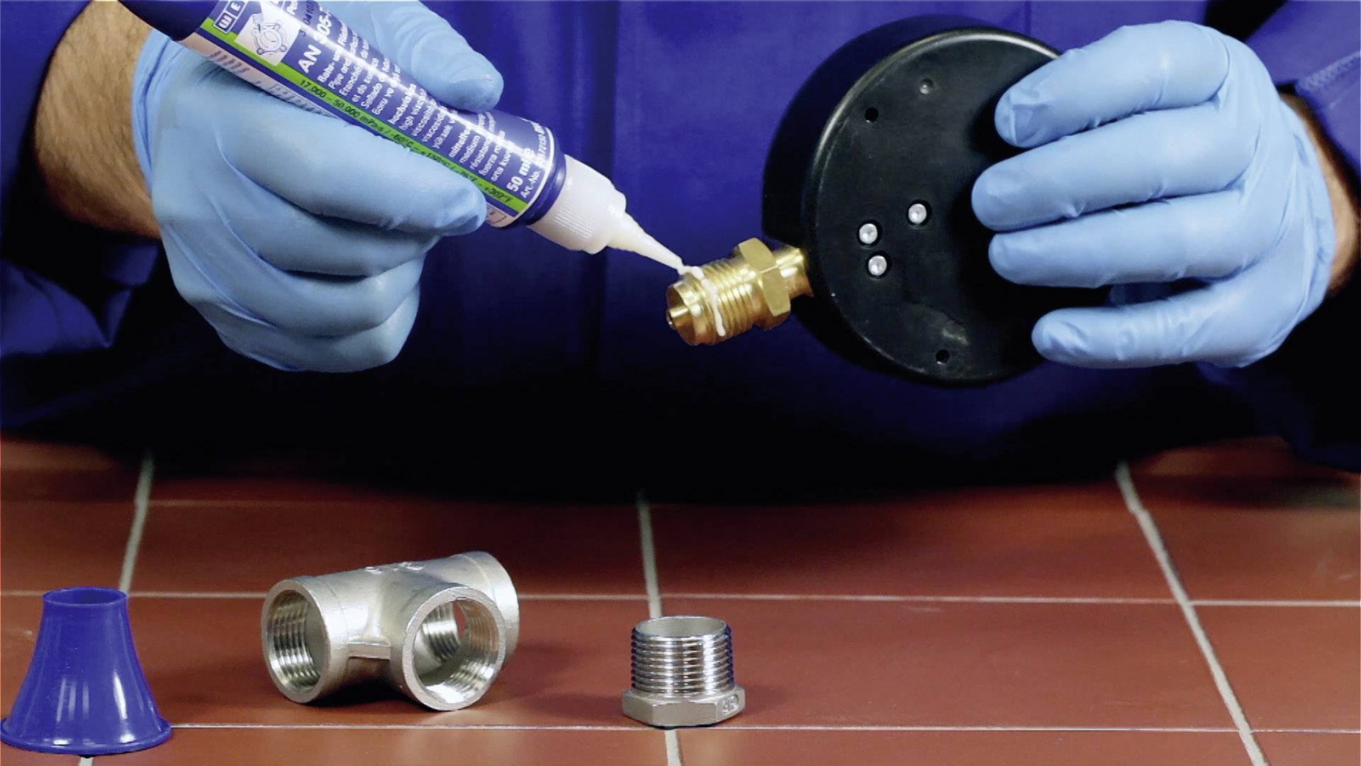 Une personne portant des gants bleus applique un produit d'étanchéité sur un filetage de tuyau en laiton. Un té et un adaptateur sont posés devant.