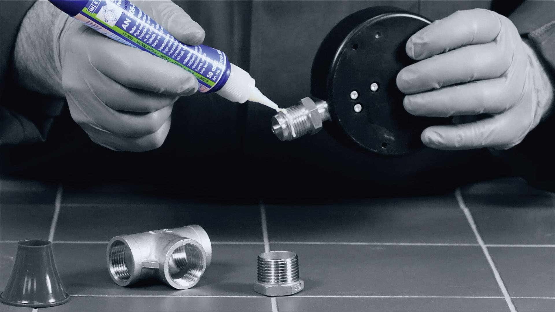 Une personne applique un agent d'étanchéité sur un filetage métallique. Au premier plan, d'autres connexions métalliques sont posées sur une table.