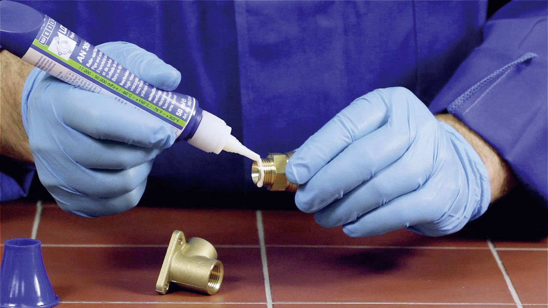 Une personne portant des gants bleus applique de la colle sur un filetage métallique afin de le connecter à une autre pièce.