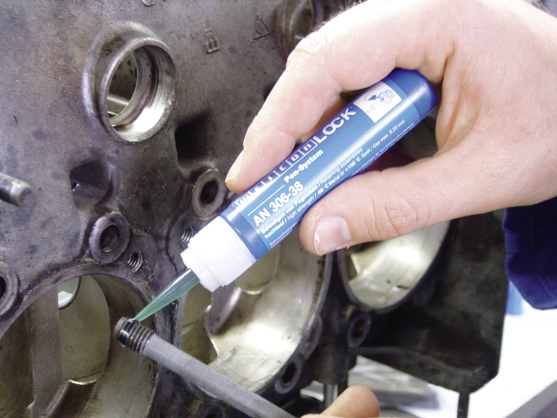 Une main applique un liquide bleu provenant d'un tube sur une vis dans une pièce métallique, probablement pour la fixation.