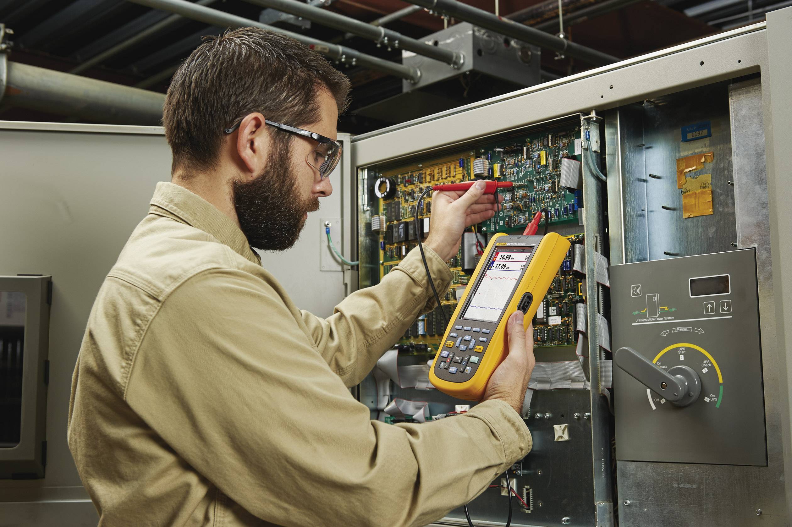 Un technicien portant des lunettes de protection utilise un multimètre pour la maintenance d'un tableau électrique dans un environnement industriel.