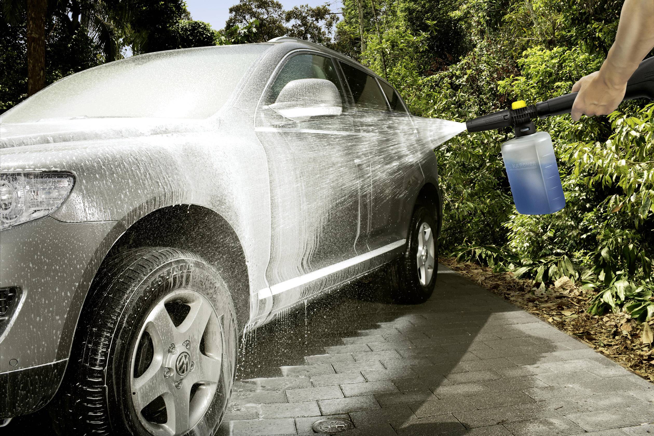 Une voiture est lavée à l'extérieur avec un nettoyeur haute pression. De la mousse recouvre la surface, tandis que l'eau scintille au soleil.