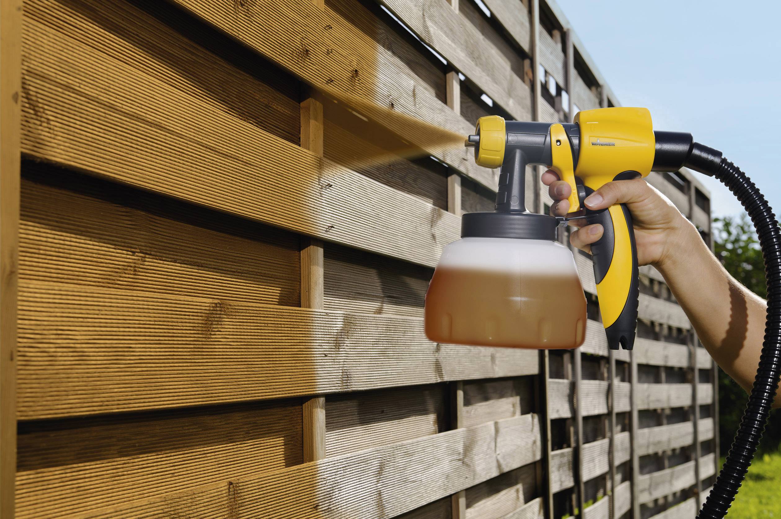 Une main pulvérise un produit de protection du bois sur une clôture de jardin en bois à l'aide d'un pistolet pulvérisateur par temps ensoleillé.