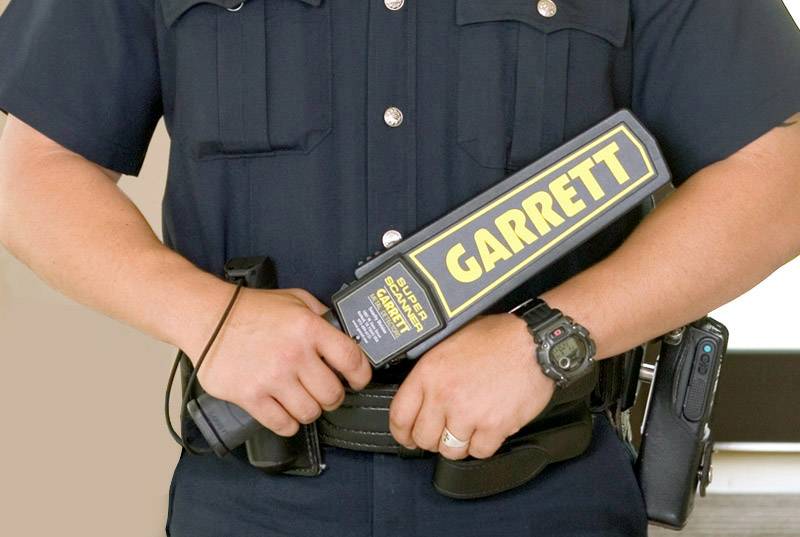 Un agent de sécurité tient un détecteur de métaux portable sur lequel est inscrit 'GARRETT'. Il porte un uniforme noir avec des accessoires de ceinture.