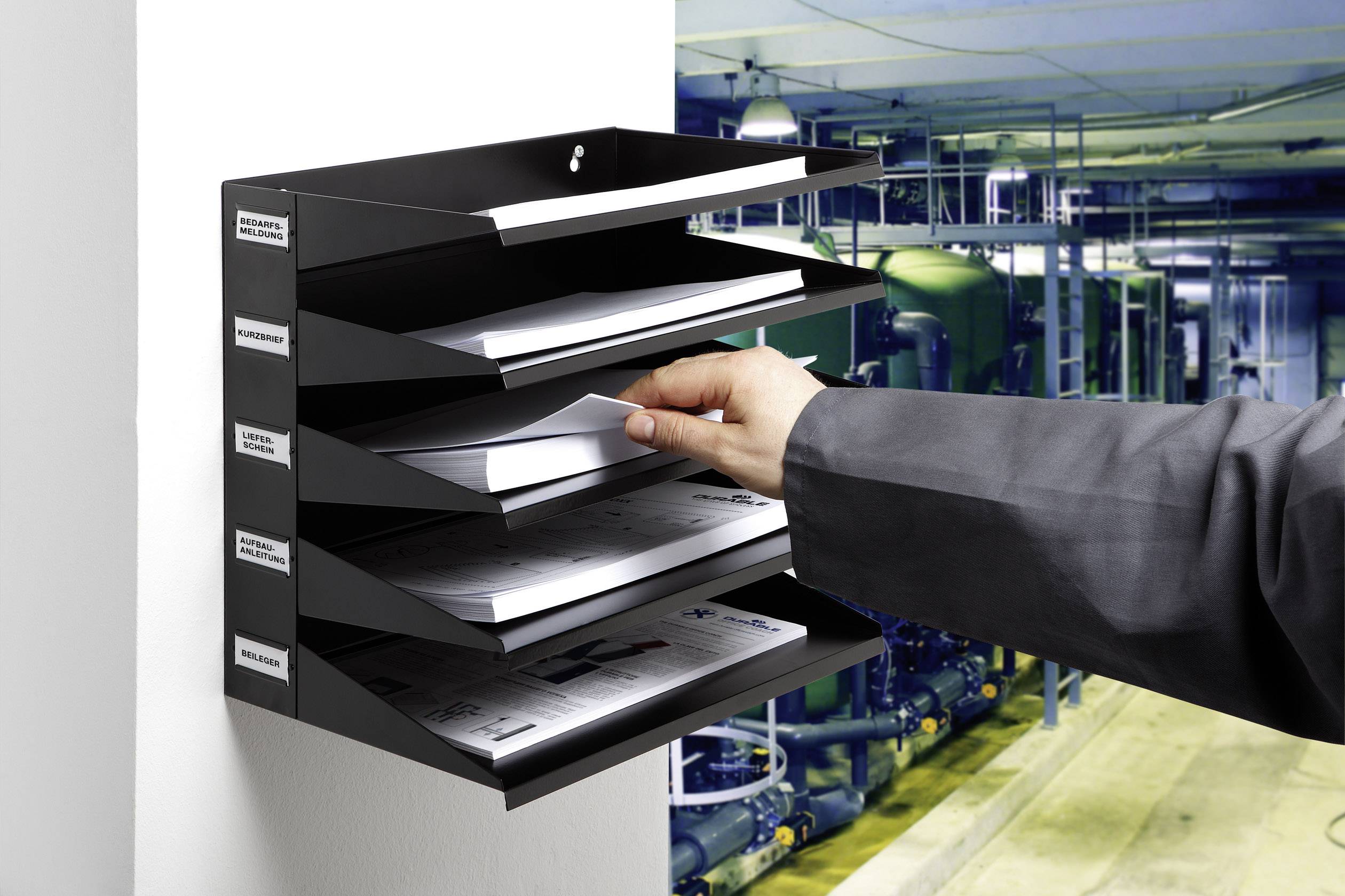 Une personne range des documents dans un système de rangement mural noir à plusieurs compartiments. En arrière-plan, on aperçoit une installation industrielle.