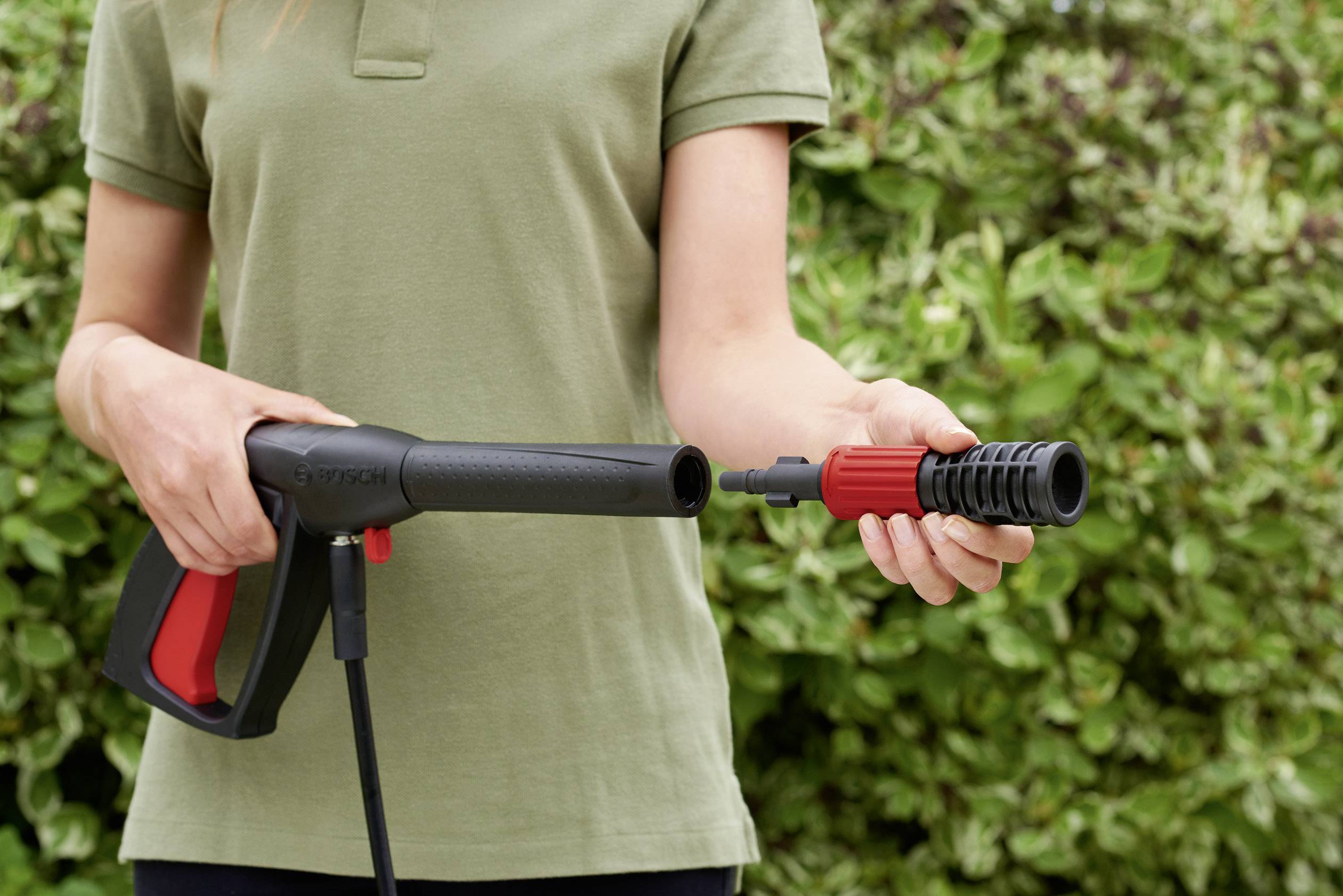Une personne montre comment raccorder une buse à un nettoyeur haute pression, sur un fond de buissons verts.