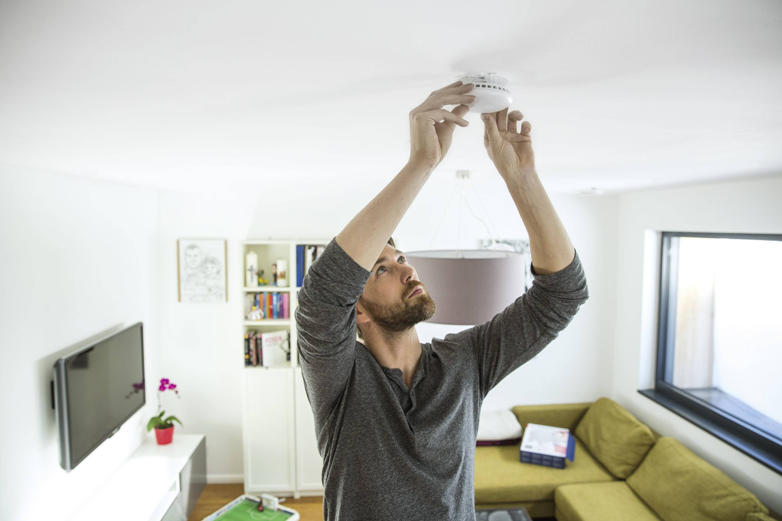 Un homme est debout dans un salon et installe un détecteur de fumée au plafond. Des meubles et une fenêtre sont visibles en arrière-plan.