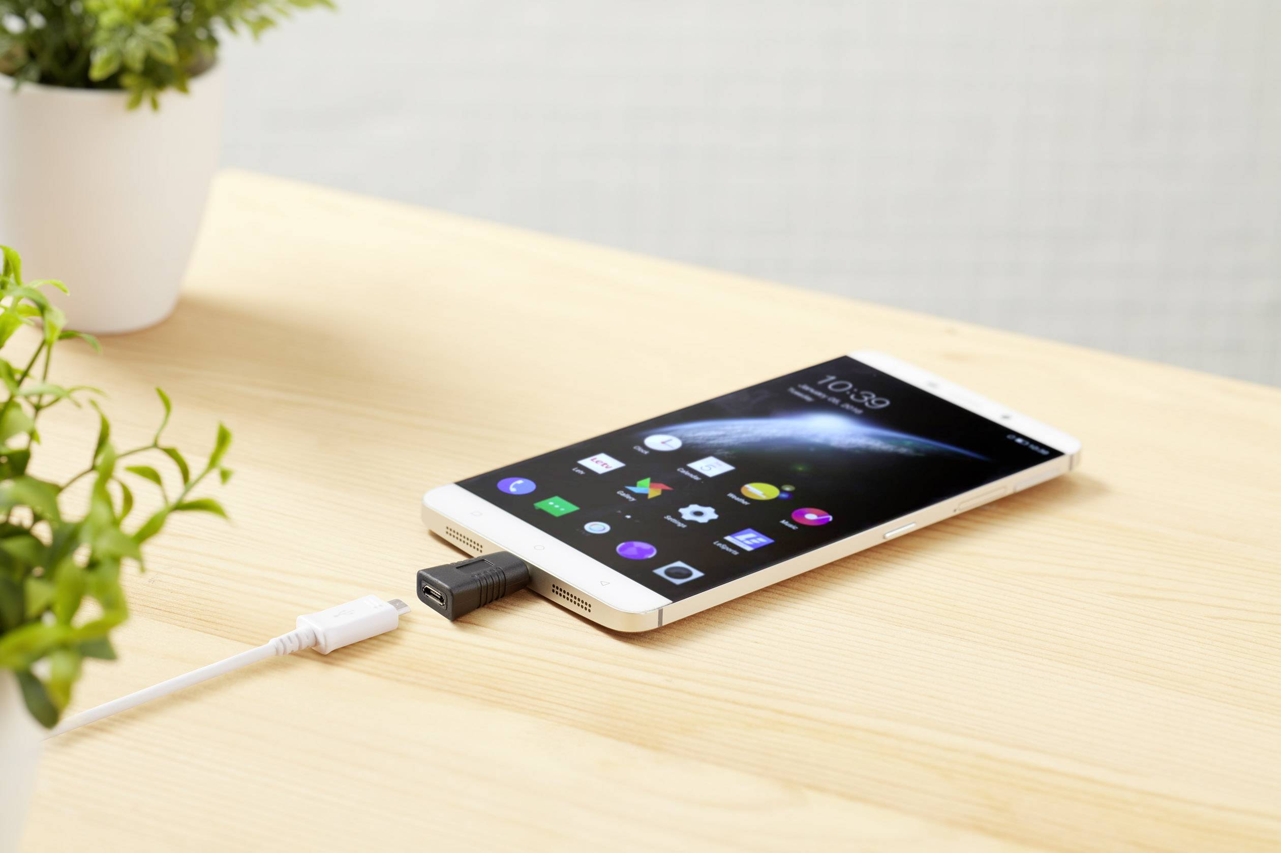 Un smartphone est posé sur une table en bois, relié par un câble USB. L'écran affiche des applications et l'heure. Des plantes sont visibles en arrière-plan.