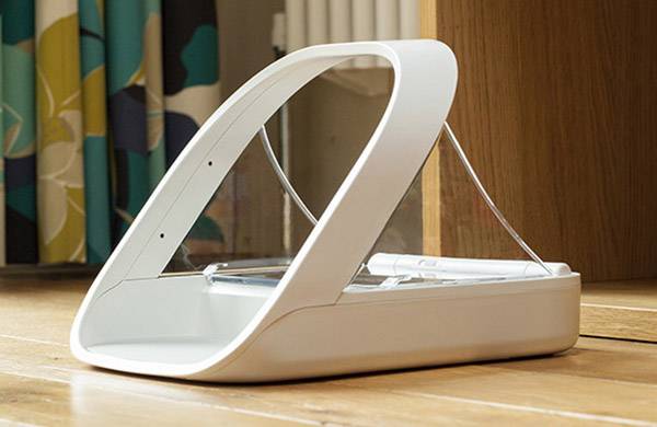 Distributeur automatique blanc pour animaux de compagnie placé sur un sol en bois, devant un rideau et un meuble en bois.