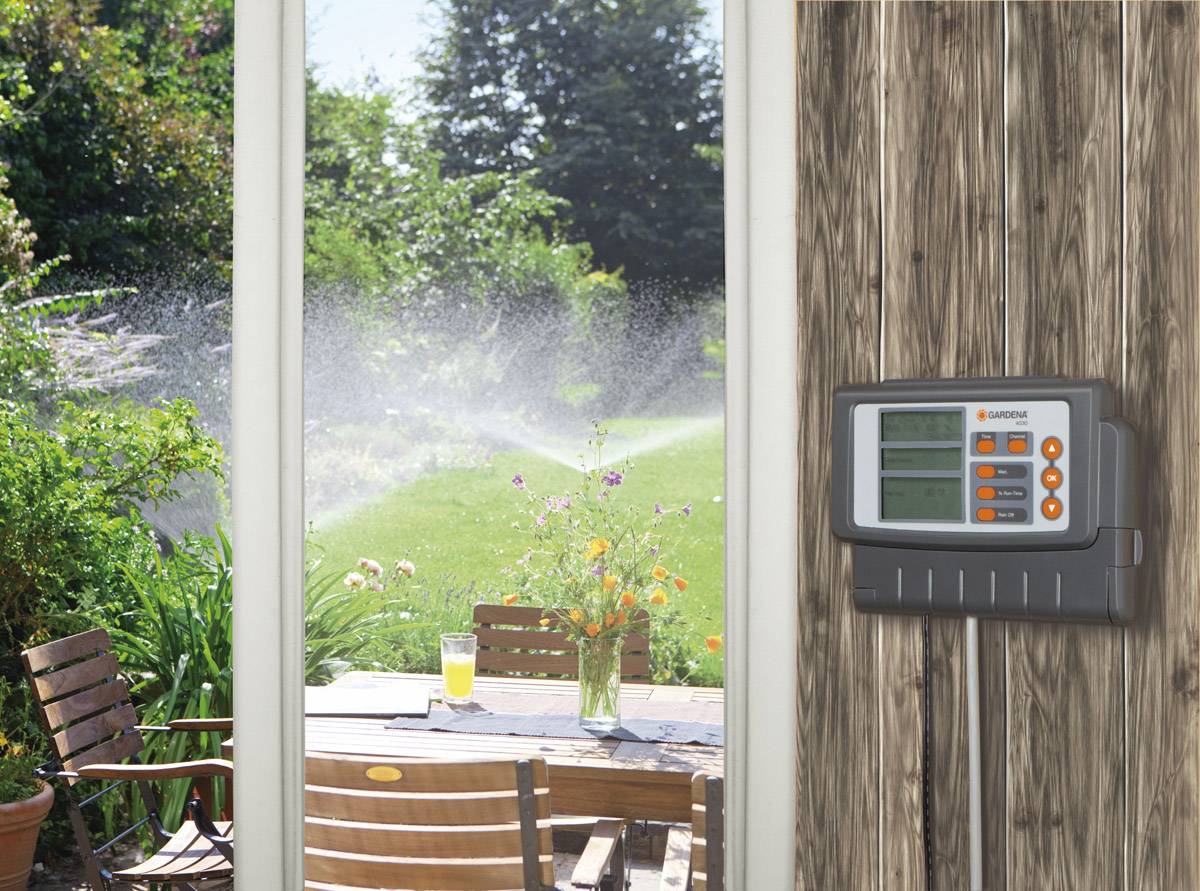 Un dispositif de commande d'irrigation est accroché à un mur en bois, donnant sur un jardin. Dans le jardin, de l'eau est pulvérisée, et une table est installée à l'extérieur.