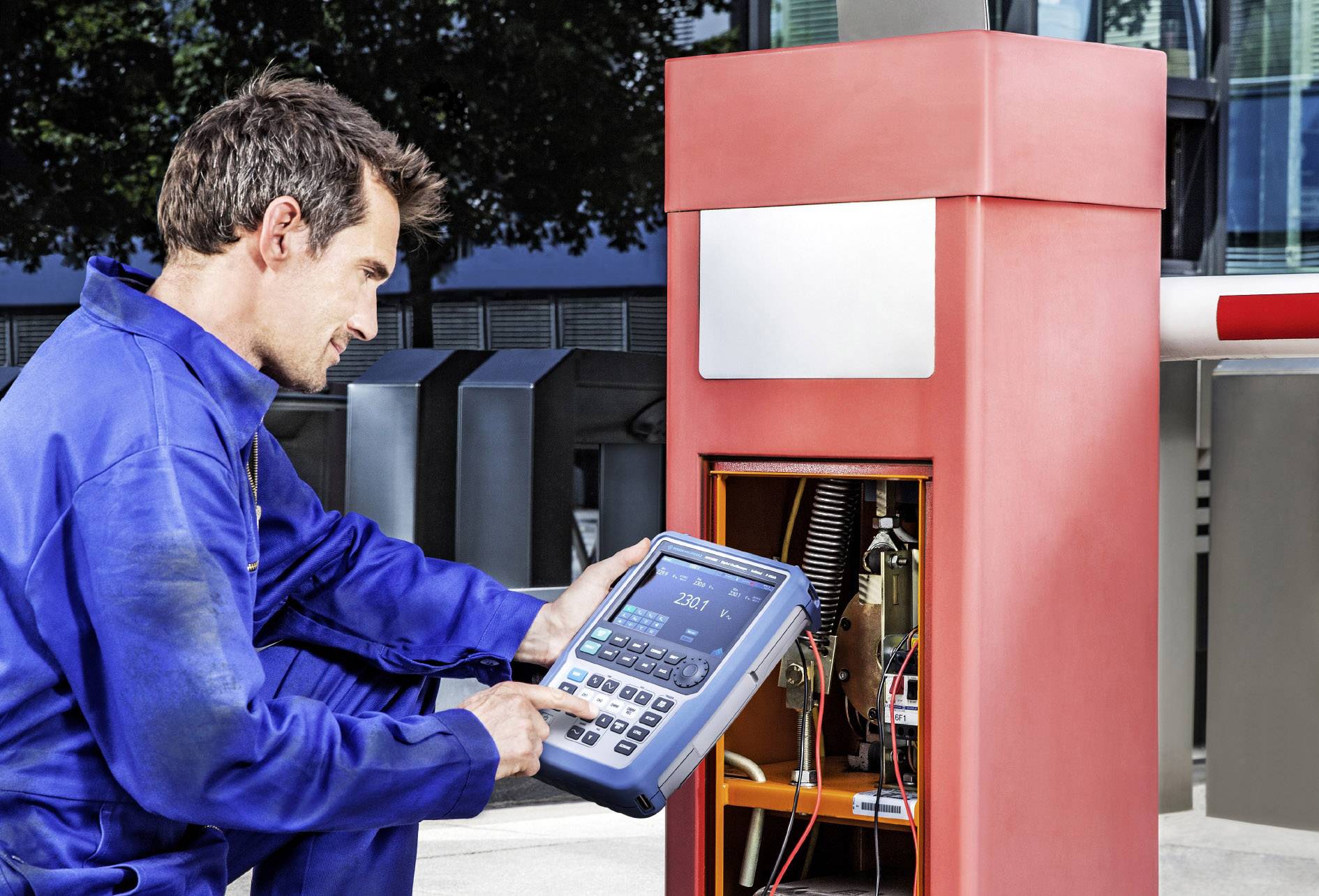 Un technicien en vêtements de travail bleus vérifie l'électronique d'une barrière rouge à l'aide d'un appareil.