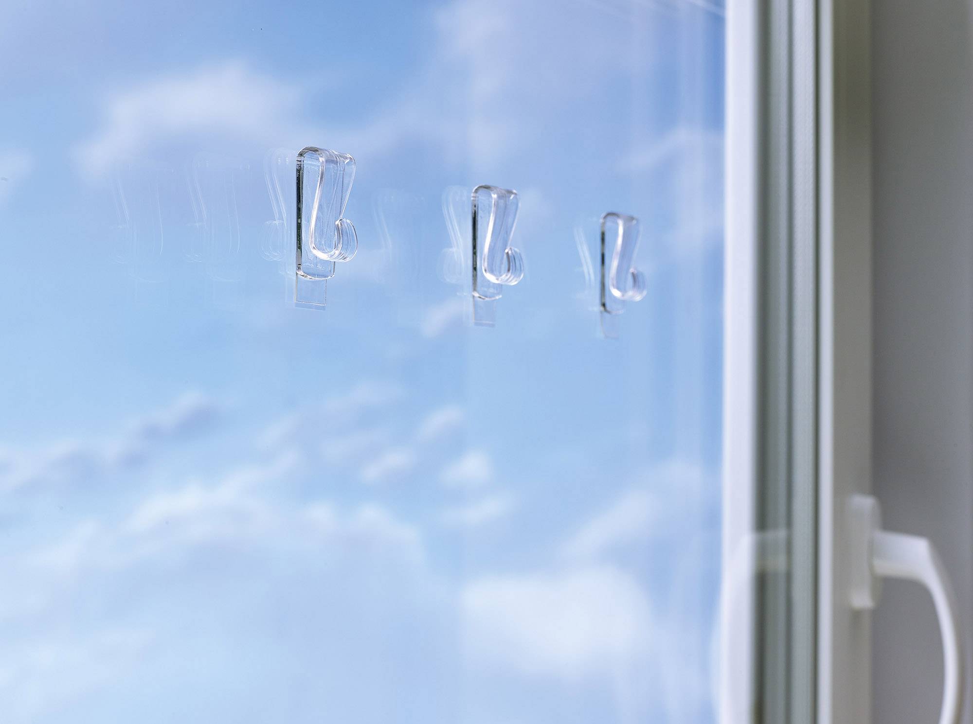 Trois crochets à vêtements transparents sont collés sur une fenêtre avec un ciel nuageux en arrière-plan. Une poignée de fenêtre est visible à droite.