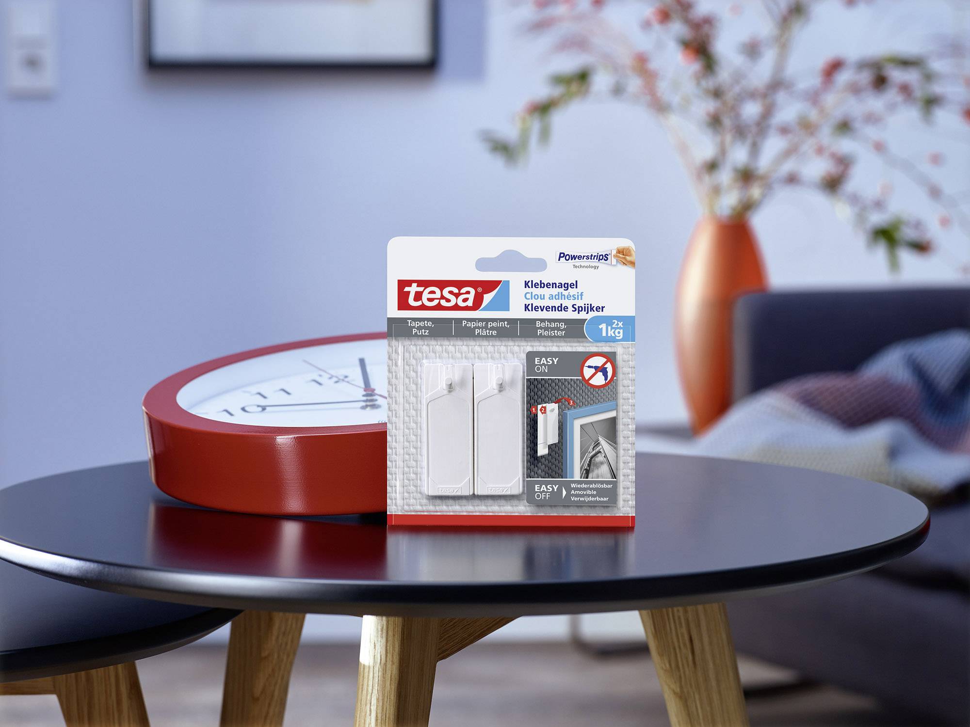 Clou adhésif tesa avec une capacité de charge de 1 kg posé sur une table dans un salon avec une horloge rouge et un vase coloré en arrière-plan.