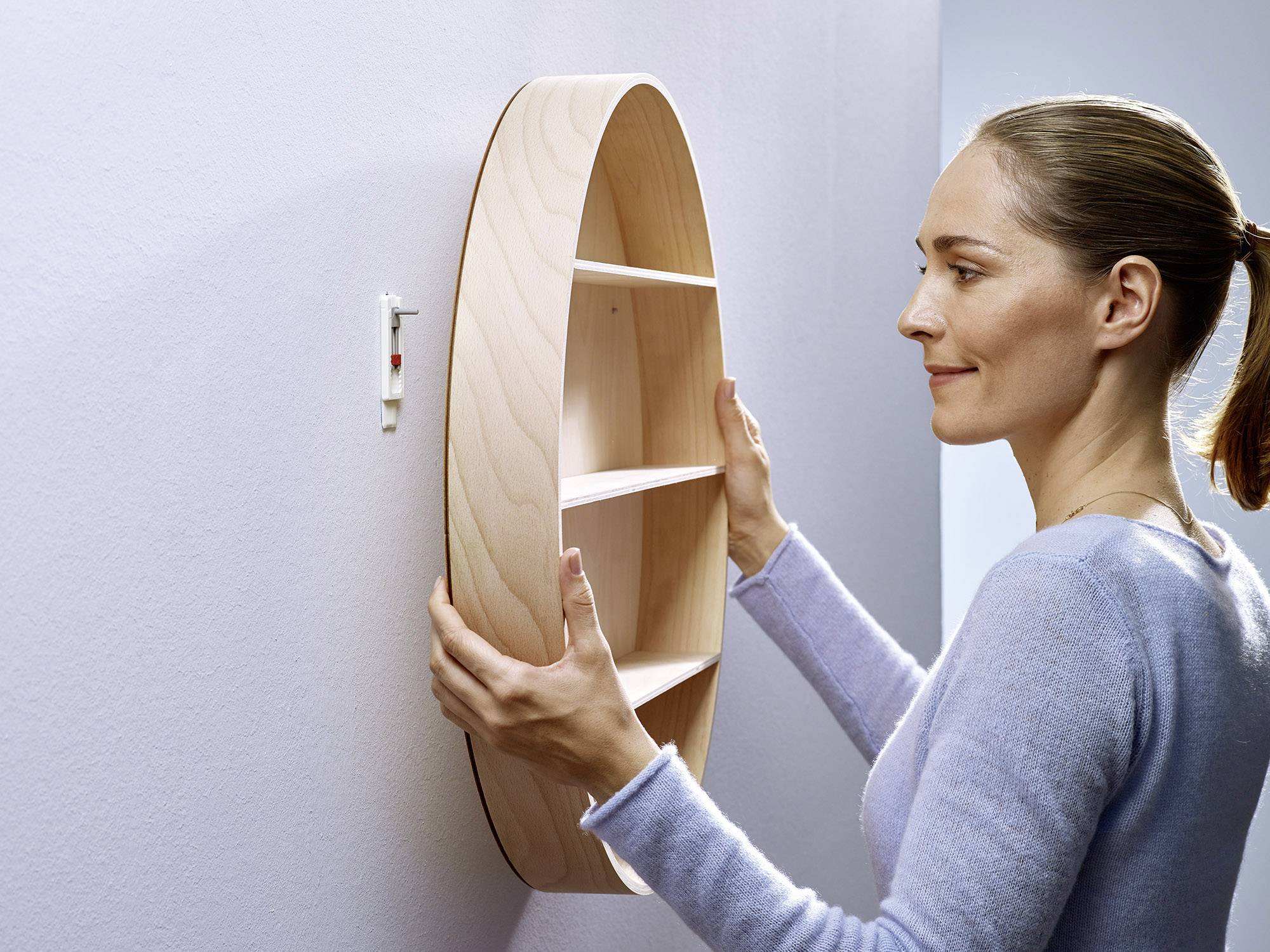 Une personne accroche une étagère ronde en bois avec plusieurs compartiments sur un mur blanc.