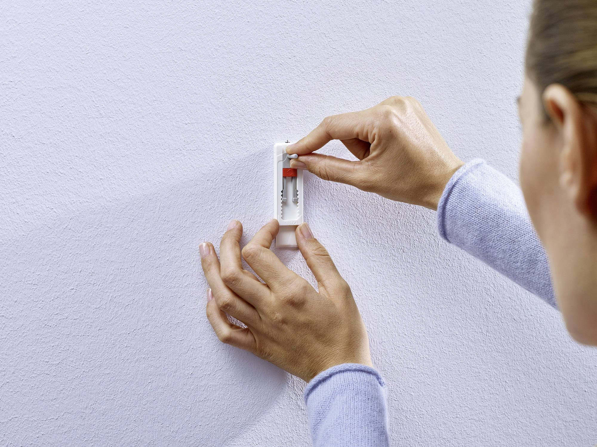 Une personne installe un petit niveau à bulle rouge et blanc sur un mur clair en utilisant ses deux mains.