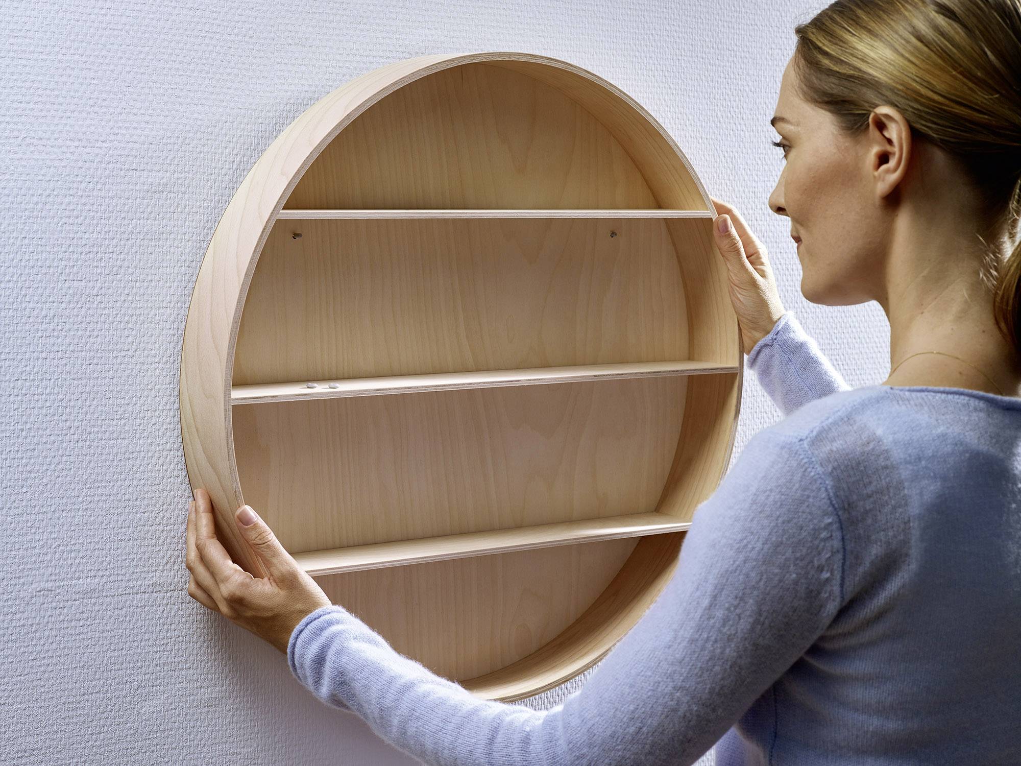 Une femme suspend une étagère ronde en bois au mur. L'étagère comporte trois niveaux et peut servir à la décoration ou au rangement.