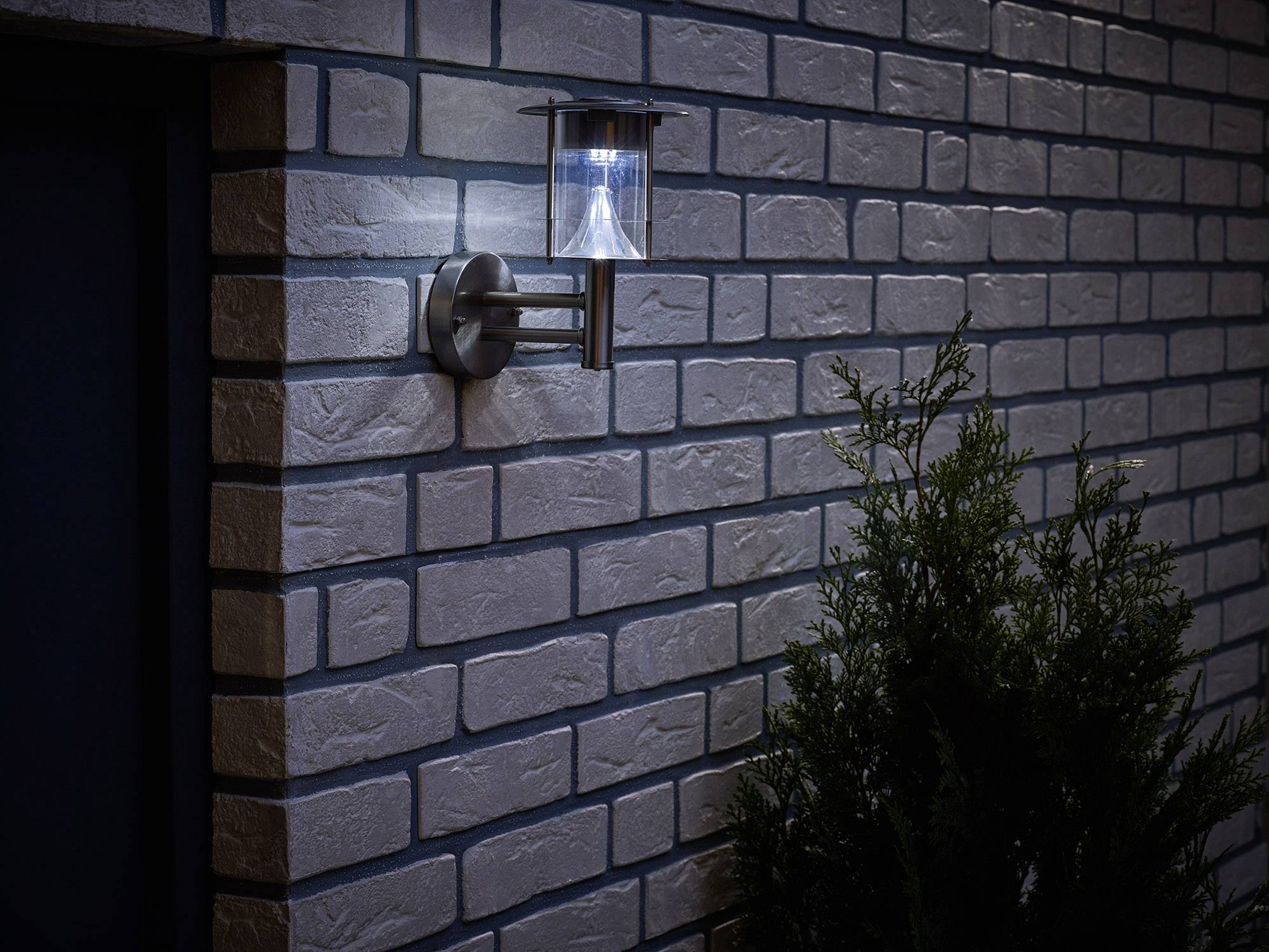 Une lampe extérieure fixée à un mur de briques brille dans l'obscurité, illuminant un buisson adjacent.