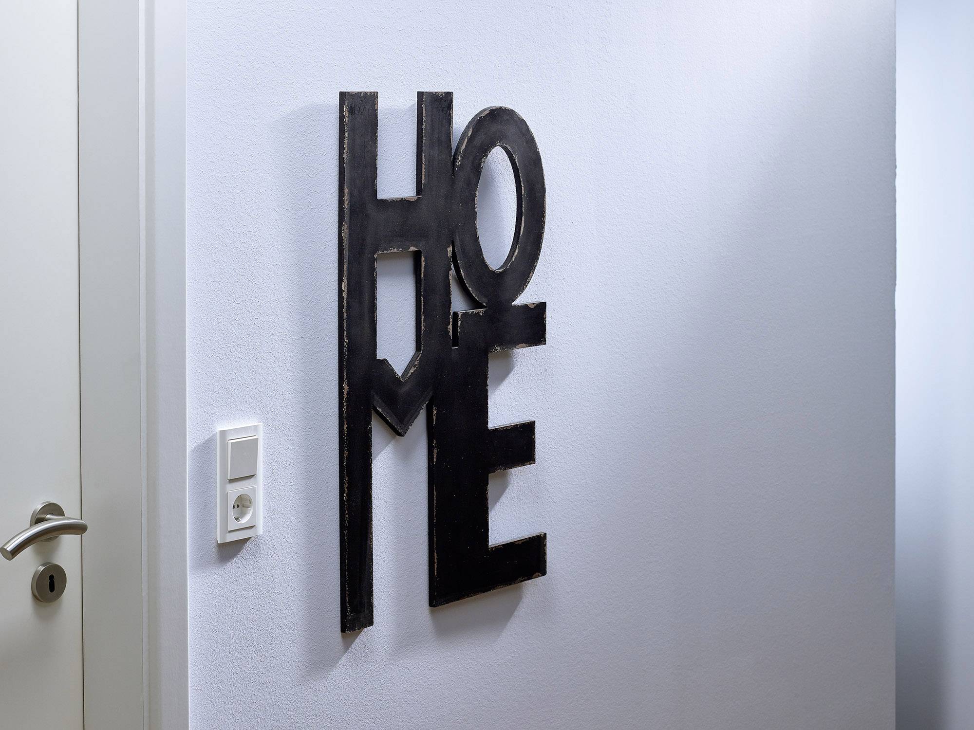 Des lettres noires sur un mur blanc forment le mot 'HOME'. Un interrupteur et une porte sont partiellement visibles à gauche.