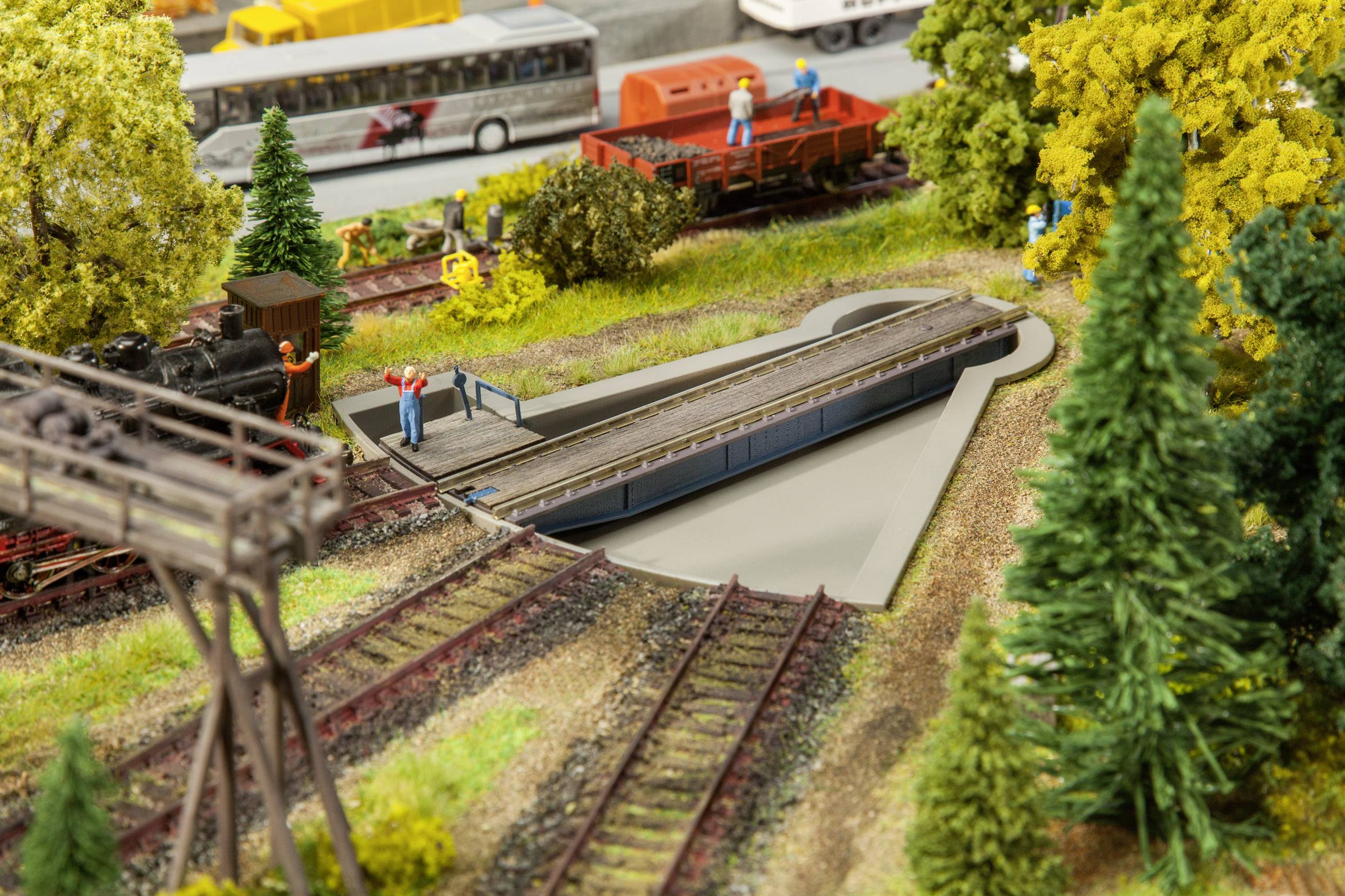 Une maquette de train miniature montre des personnages sur une plaque tournante, entourés d'arbres et de rails. Des trains sont stationnés en arrière-plan.