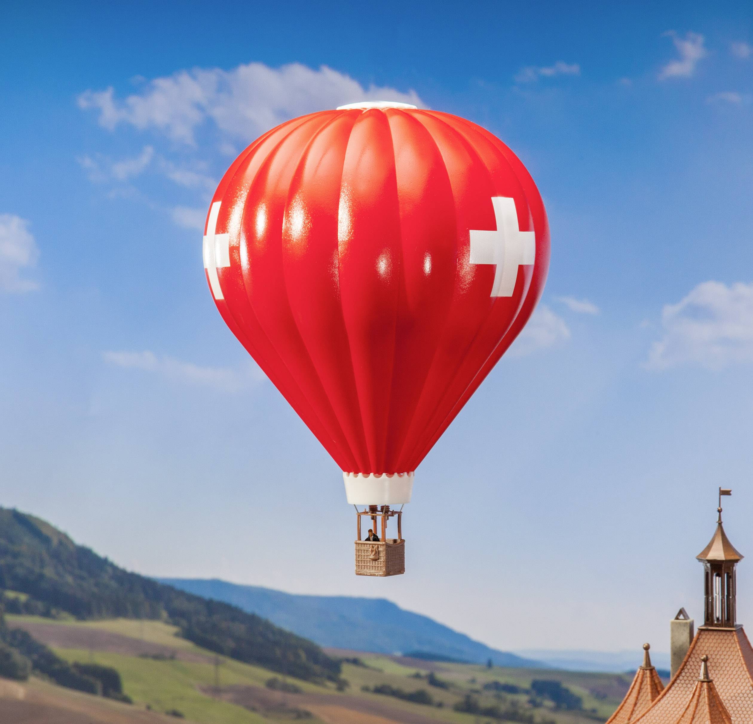 Ballon à air chaud avec des croix rouges et blanches flottant au-dessus d'un paysage vallonné avec une tour de bâtiment au premier plan.