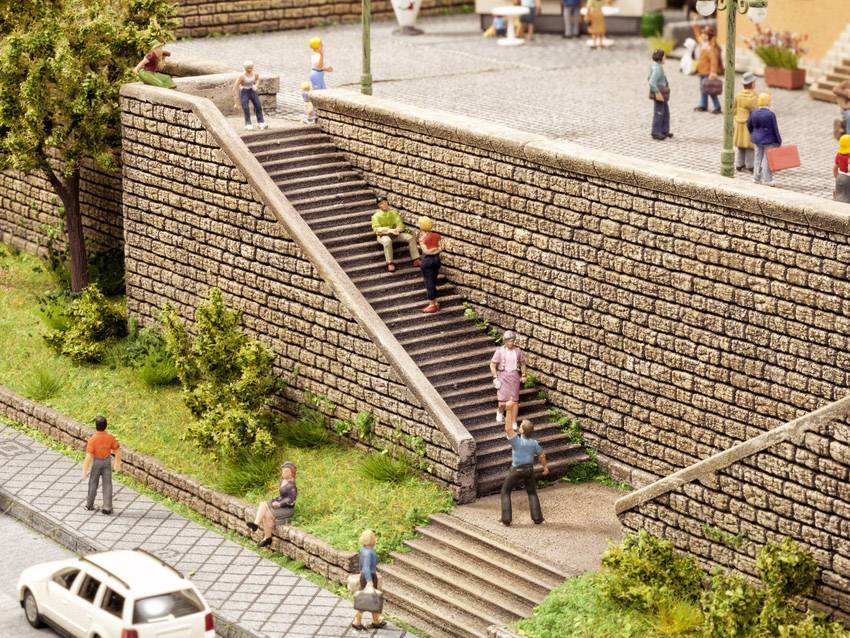 Une scène miniature détaillée montre un escalier de pierre contre un mur avec des personnes, certaines assises, d'autres montant ou descendant les marches. Au premier plan, une voiture blanche est garée sur la rue.
