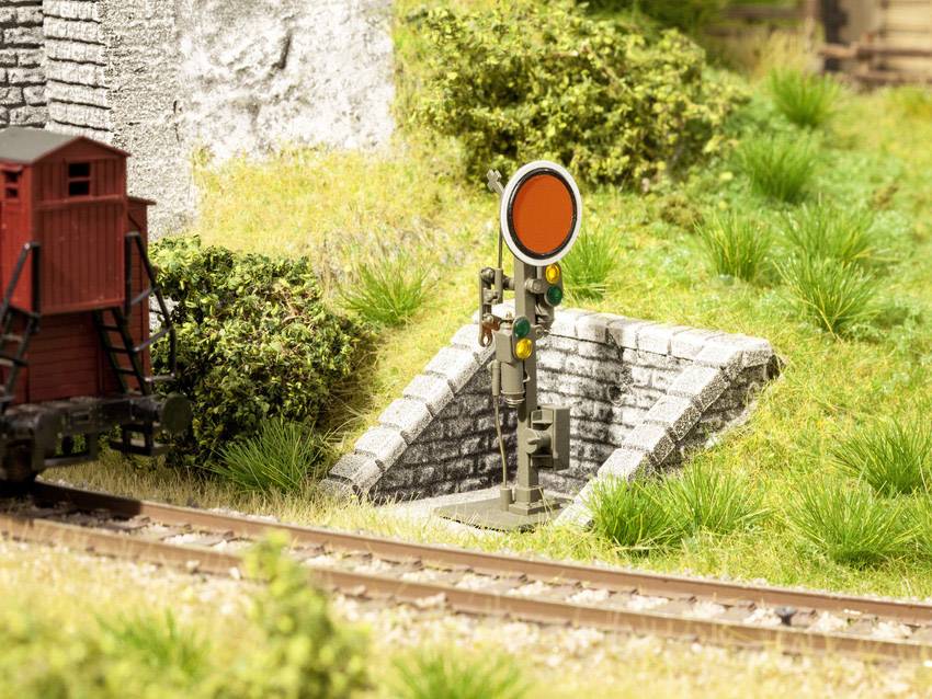 Un signal ferroviaire rouge se dresse à côté des voies ferrées dans un paysage miniature, entouré de végétation verte et d'une petite maison de briques.