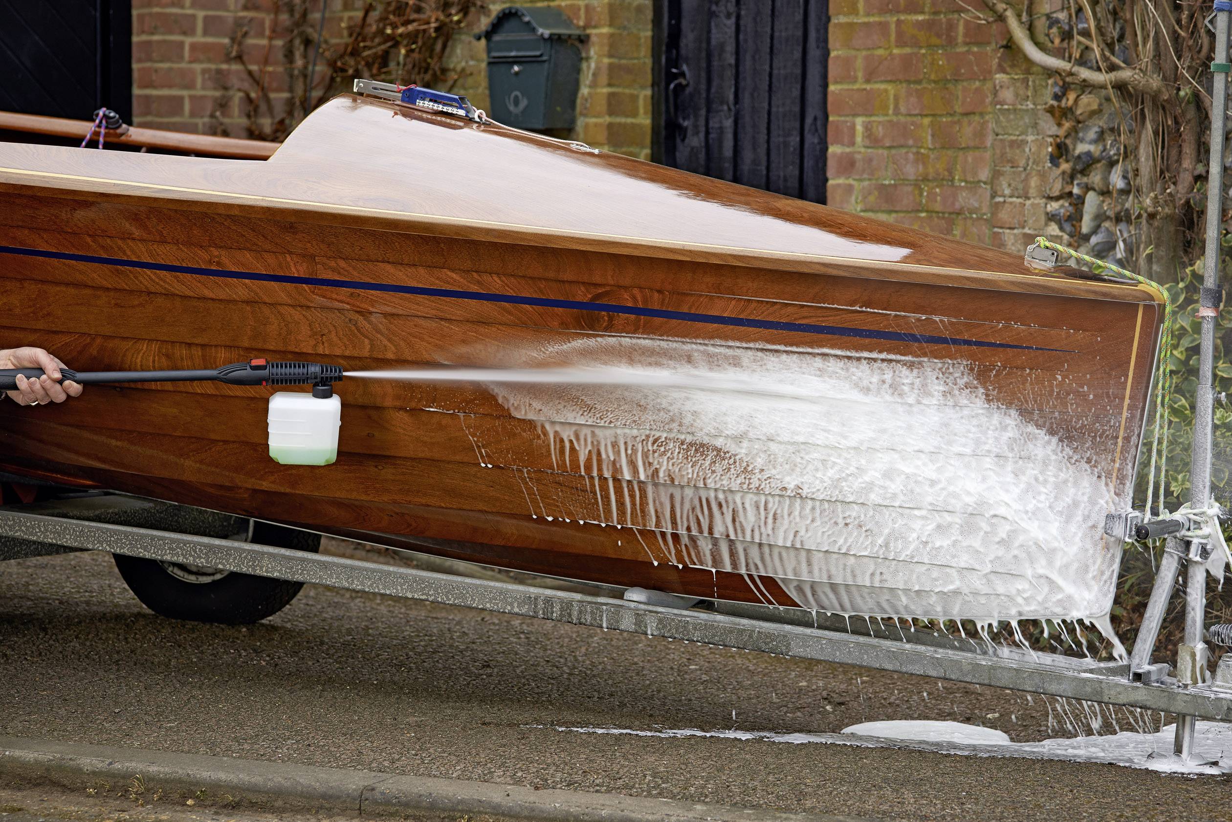 Une personne nettoie un bateau en bois à l'aide d'un nettoyeur haute pression sur une route. De la mousse recouvre partiellement la coque du bateau.