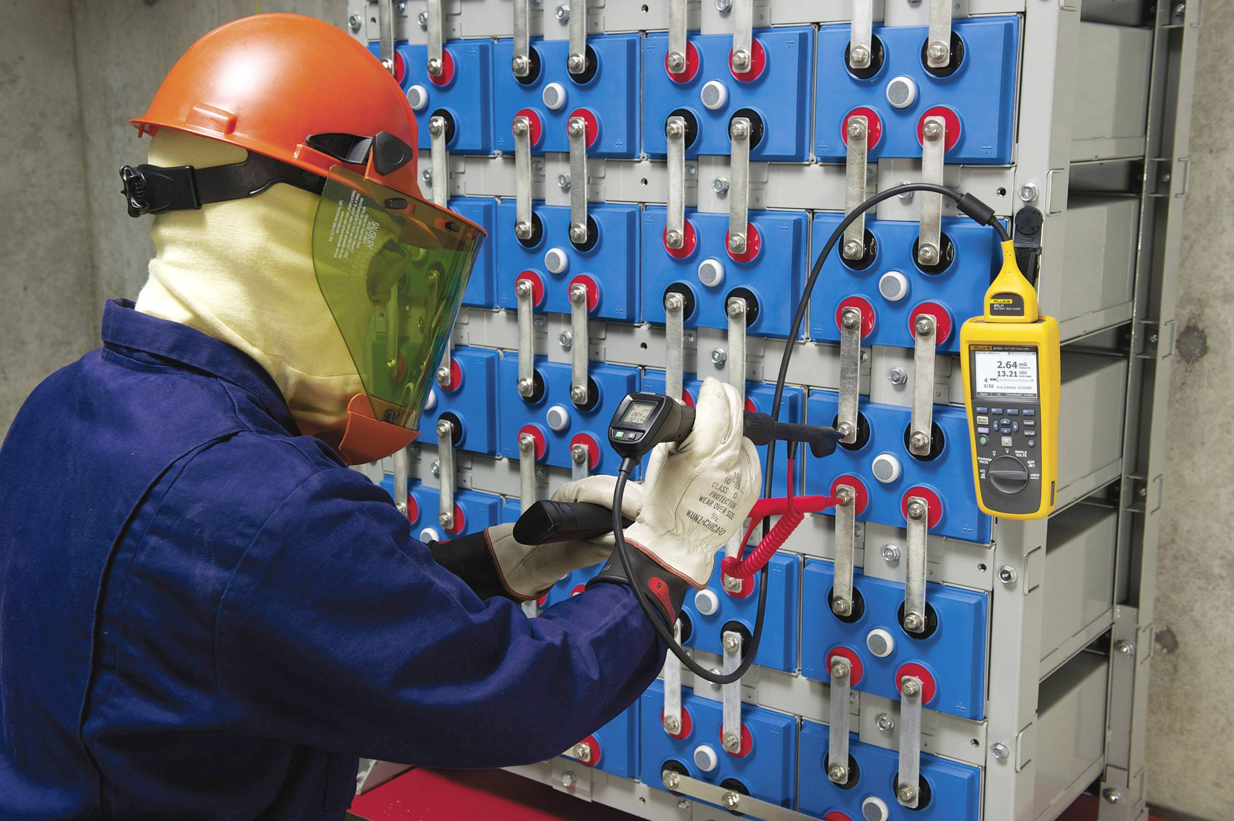 Un technicien porte un équipement de protection et vérifie la tension d'un grand système de batteries dans un environnement industriel à l'aide d'un appareil de mesure.
