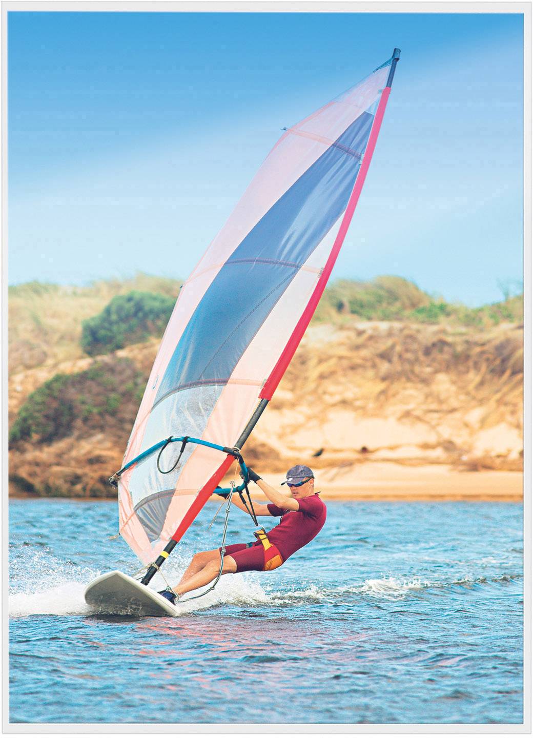 Une personne fait de la planche à voile sur l'eau, avec une voile colorée et un arrière-plan côtier boisé pittoresque.