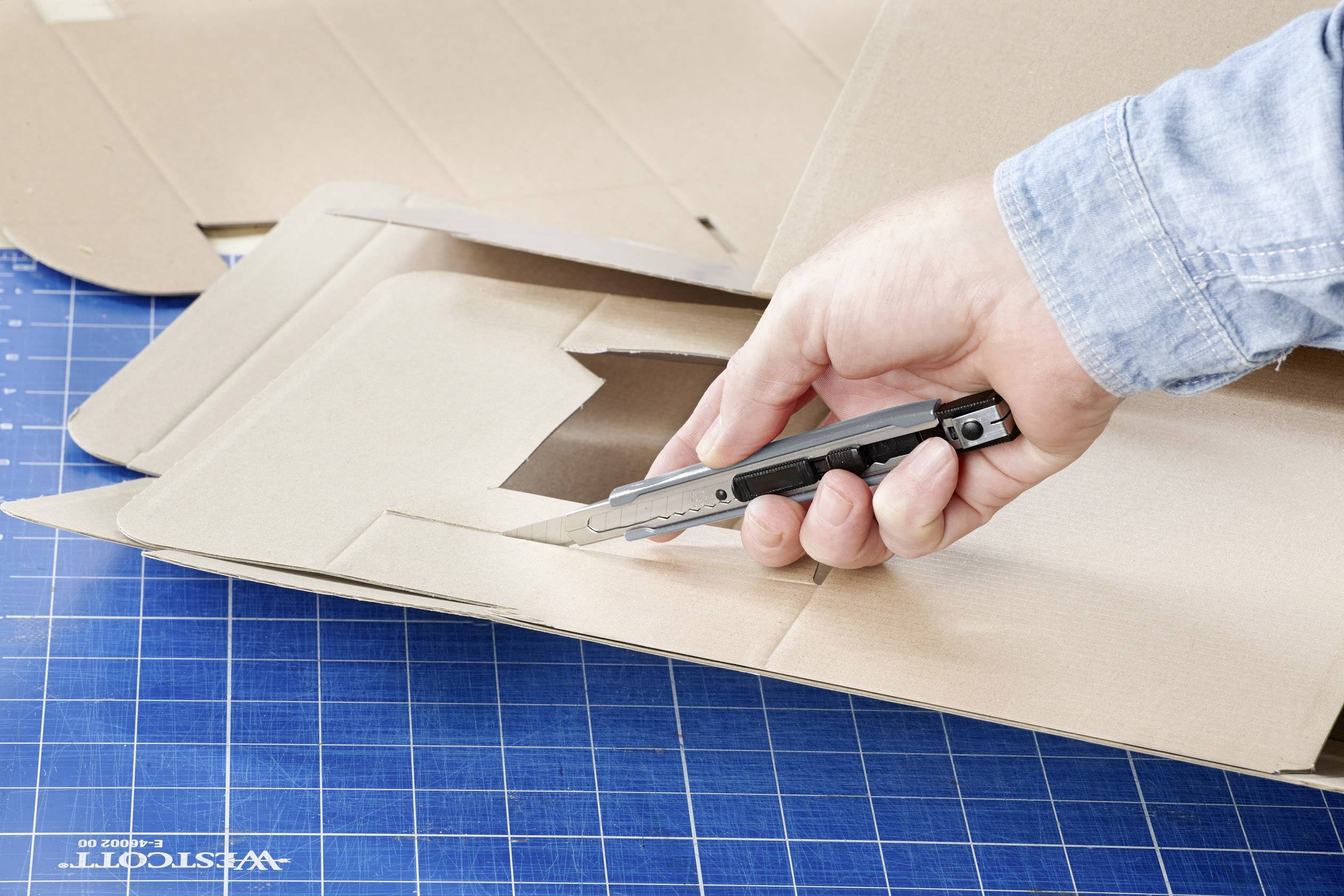Une main utilise un cutter pour découper un carton plat sur un tapis de coupe bleu quadrillé.