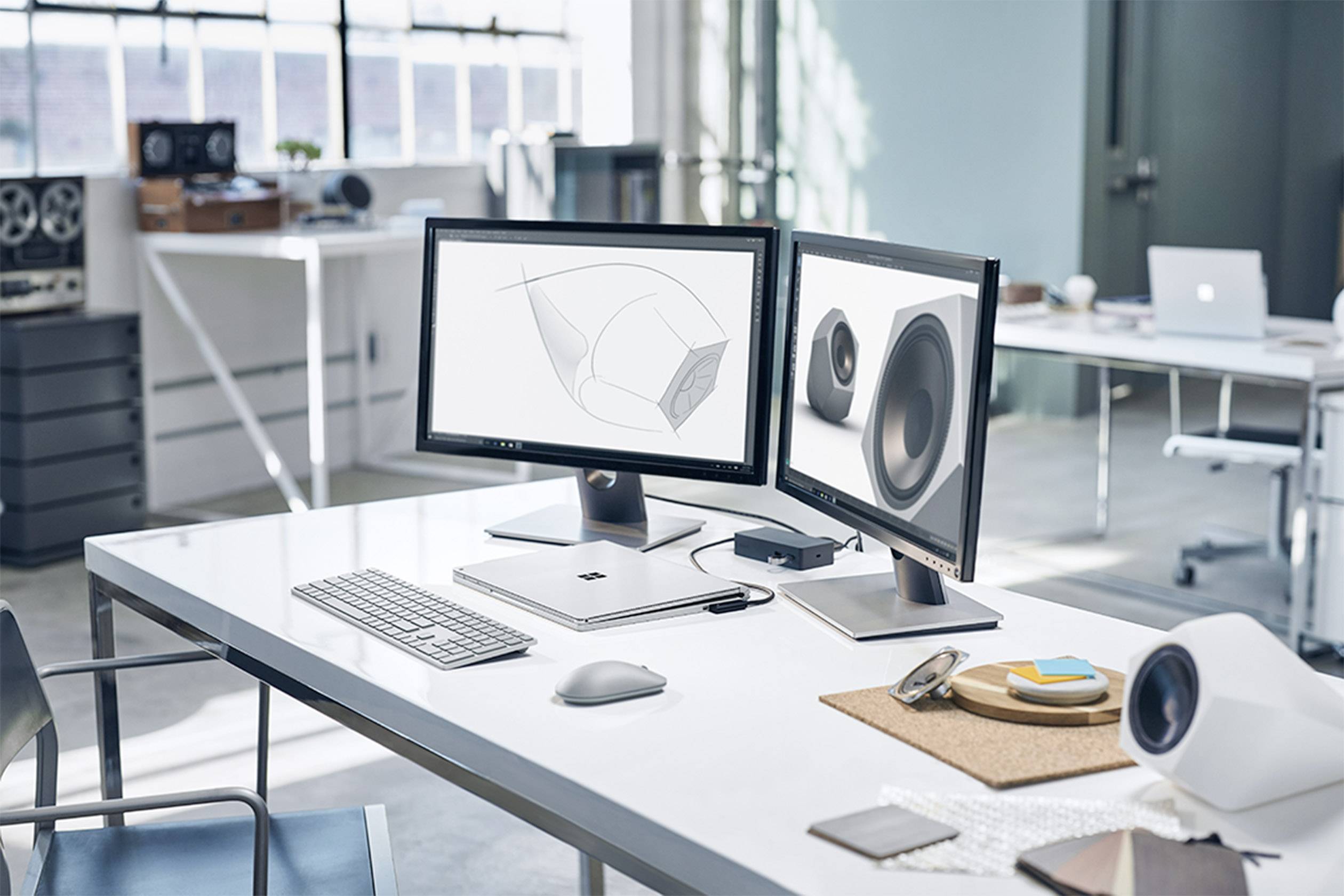 Bureau moderne avec deux écrans affichant des designs 3D. Sur une table sont disposés un clavier, une souris et des haut-parleurs. Aménagement lumineux et minimaliste.